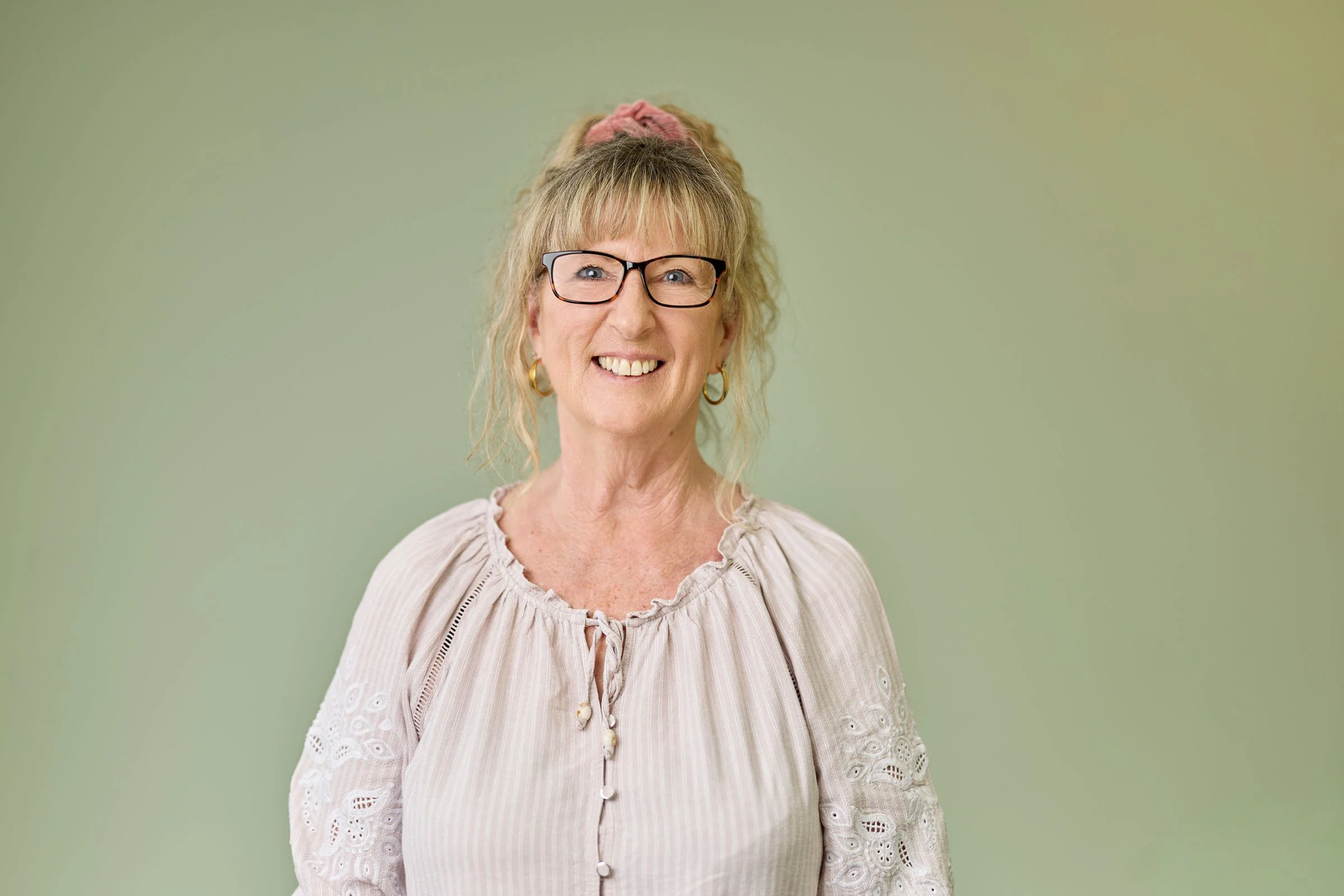 An elderly woman with blond hair, glasses, and hoop earrings smiling, wearing a light-colored blouse with eyelet details, standing against a plain green background.