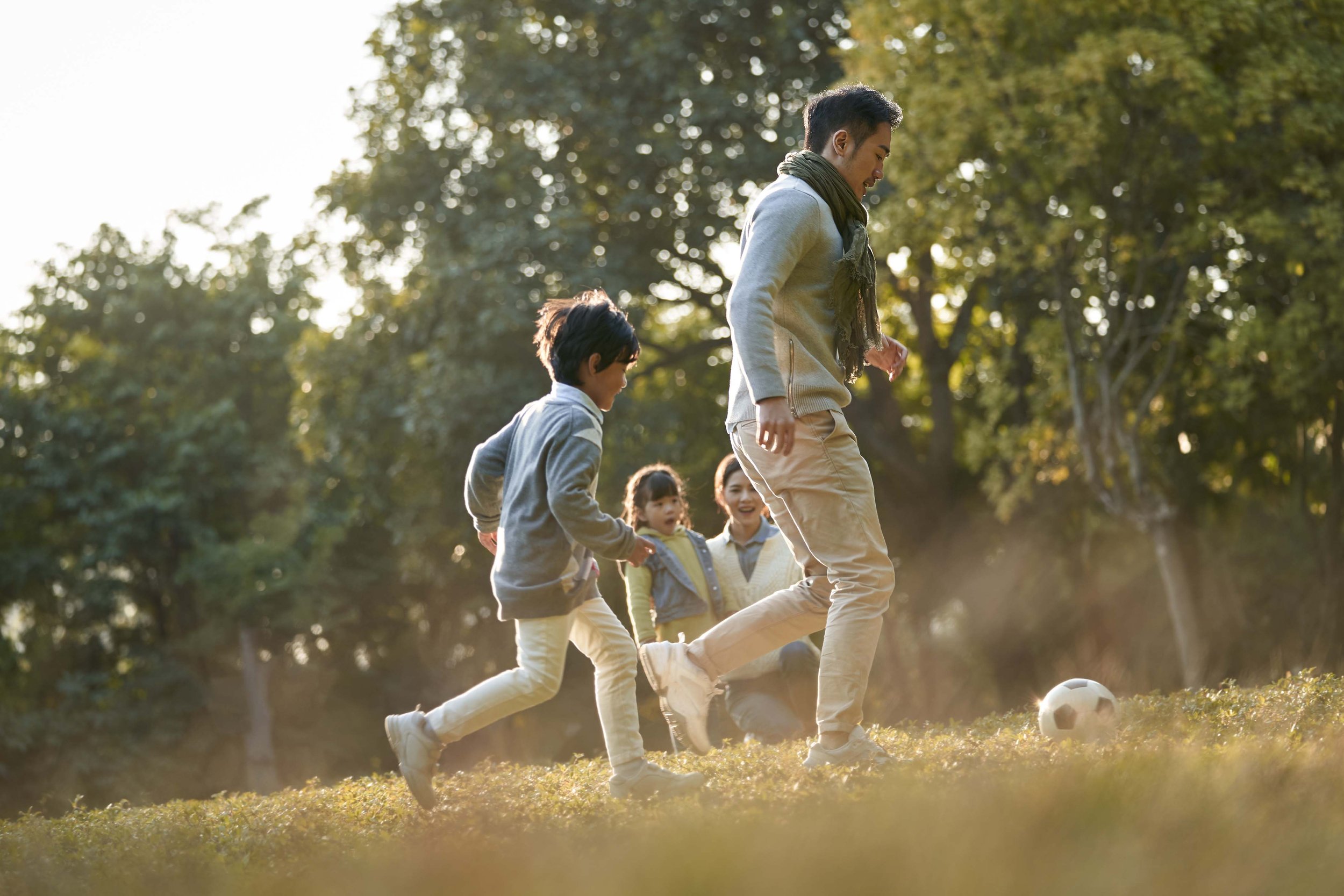 A group of children and an adult playing soccer outdoors in a park with trees, during sunset.