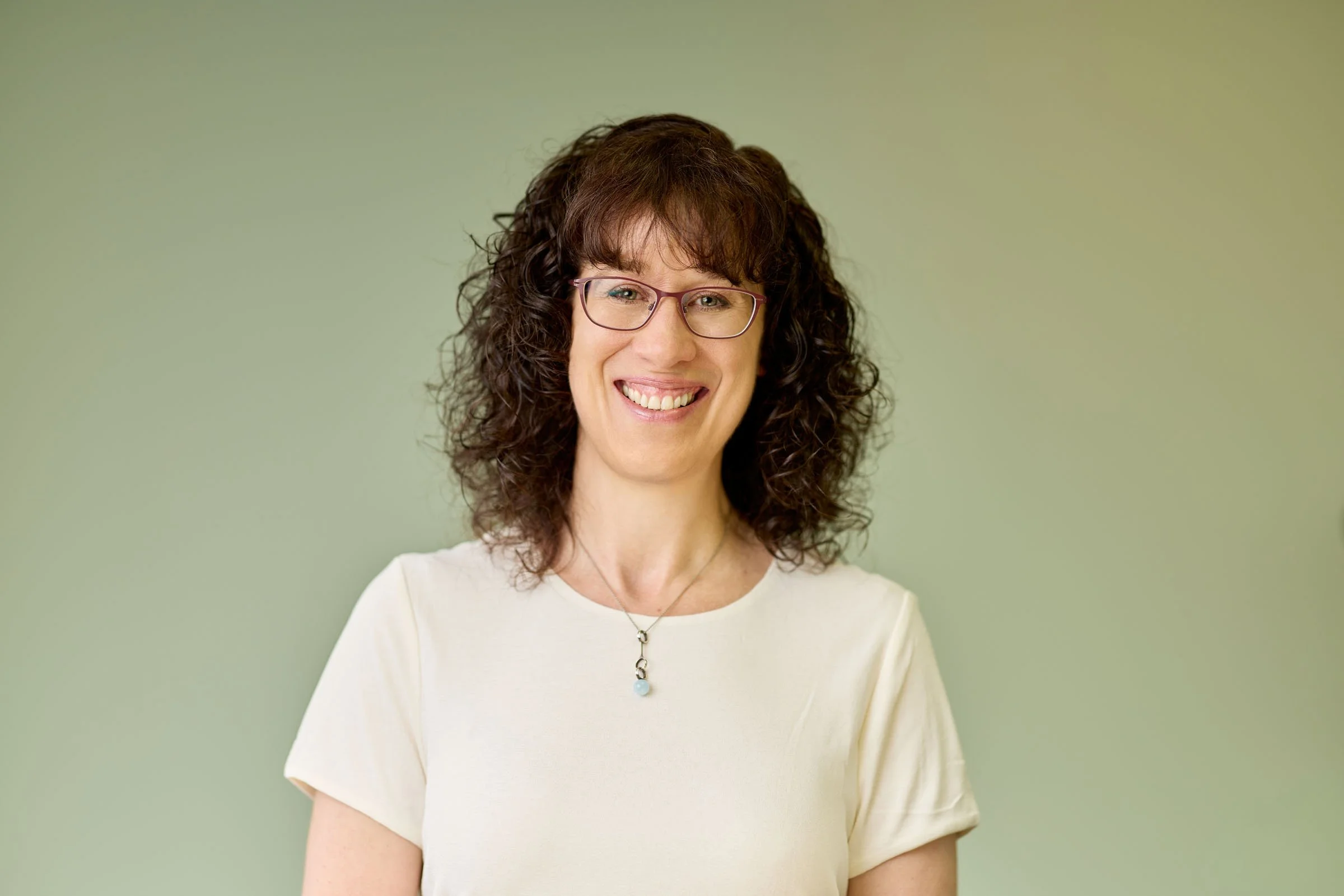 A woman with curly brown hair, wearing glasses and a white top, smiling against a light green background.