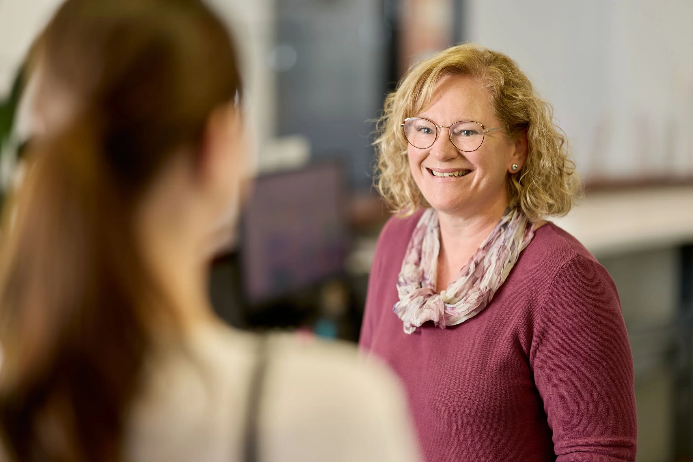 A woman with curly blonde hair, glasses, and earrings smiling while talking to another woman with brown hair, in an indoor setting.
