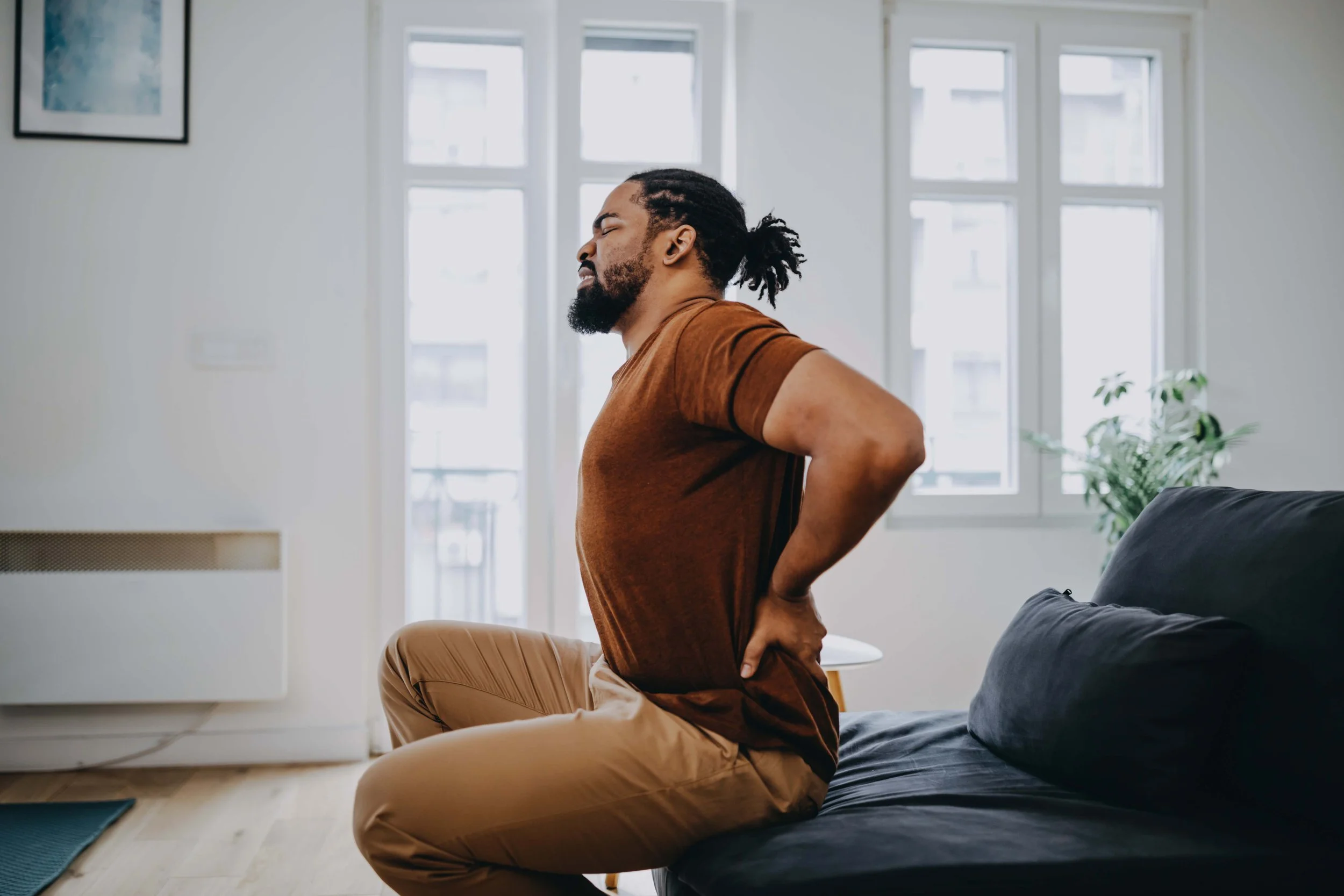 A man with dreadlocks and a beard is sitting on a sofa in a bright room, stretching his back with his eyes closed.