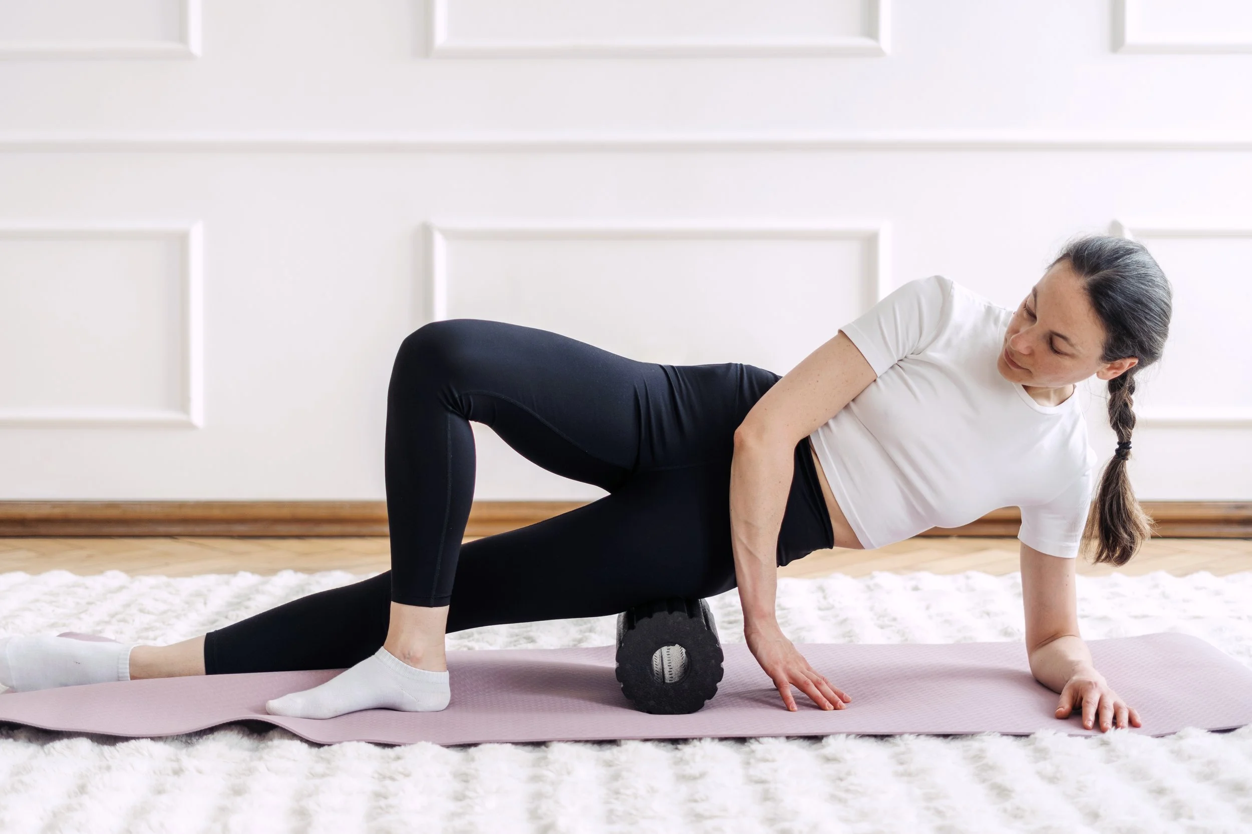 Woman lying on folded yoga mat with foam roller under her hips, performing exercise with a partner in a bright room with white walls.