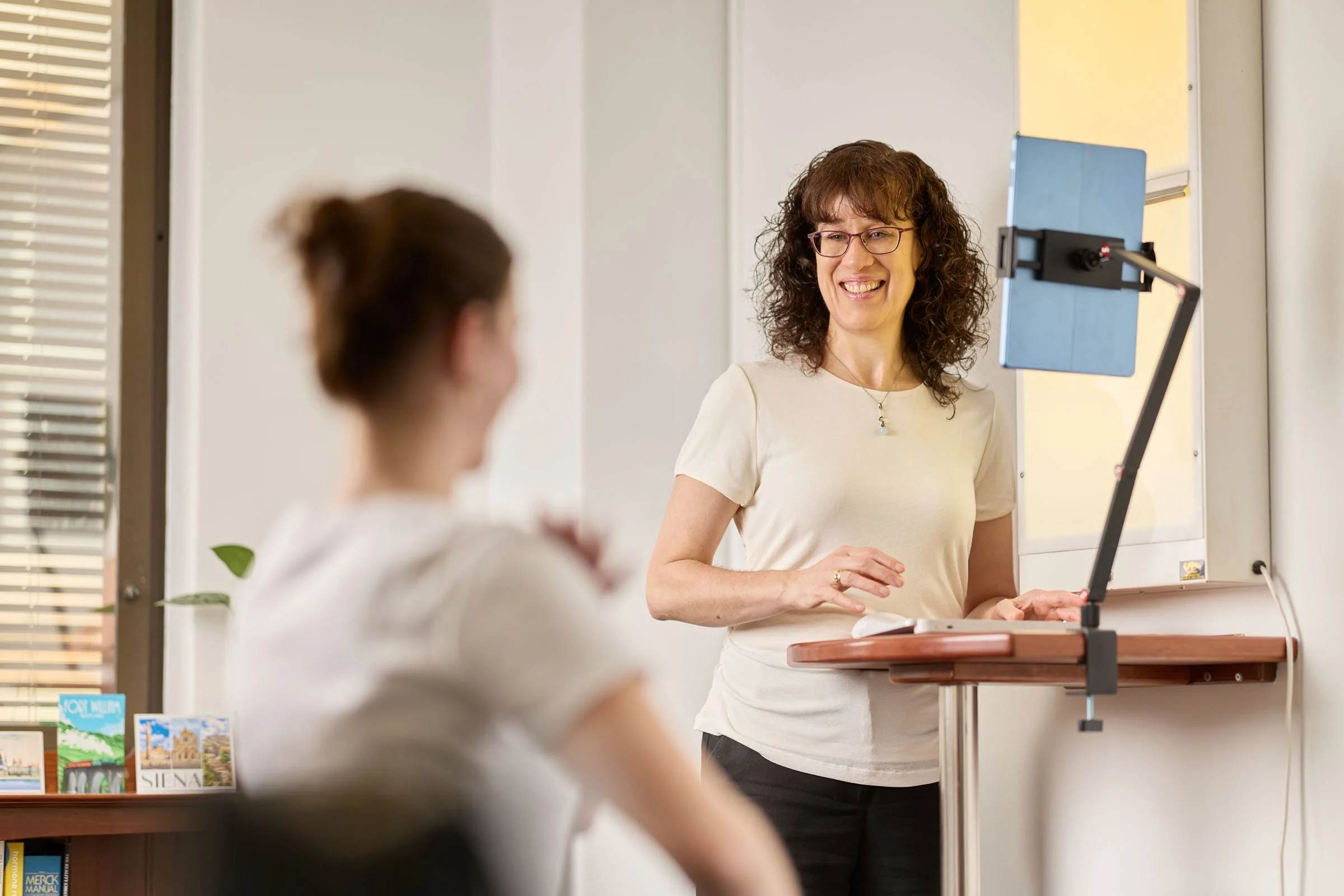 A woman with glasses and curly hair smiles while speaking to a person in a white shirt in a room with bookshelves and a window.