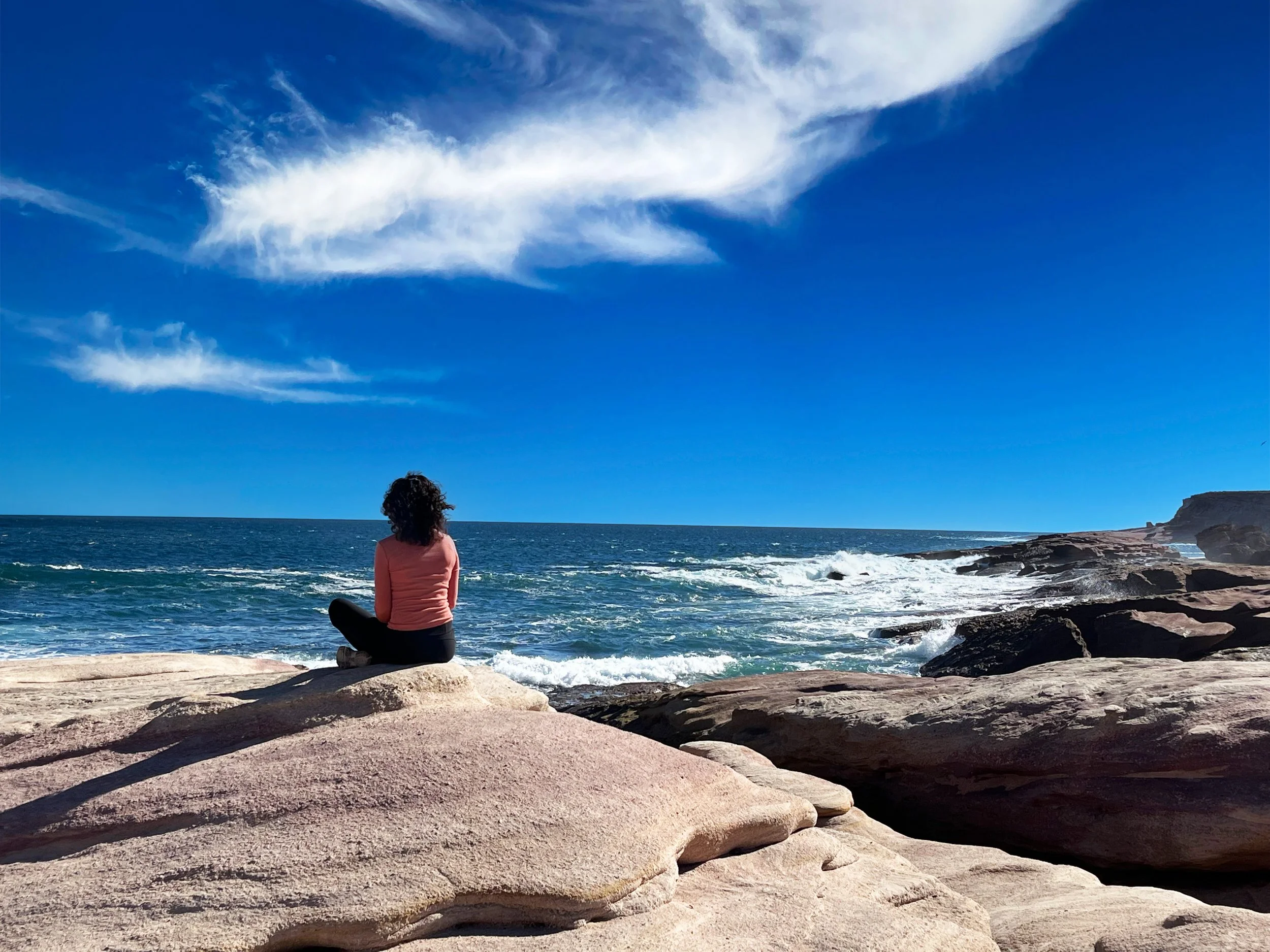 A woman sitting on rocks by the ocean, looking at the water and sky, with clouds and cliffs in the distance.