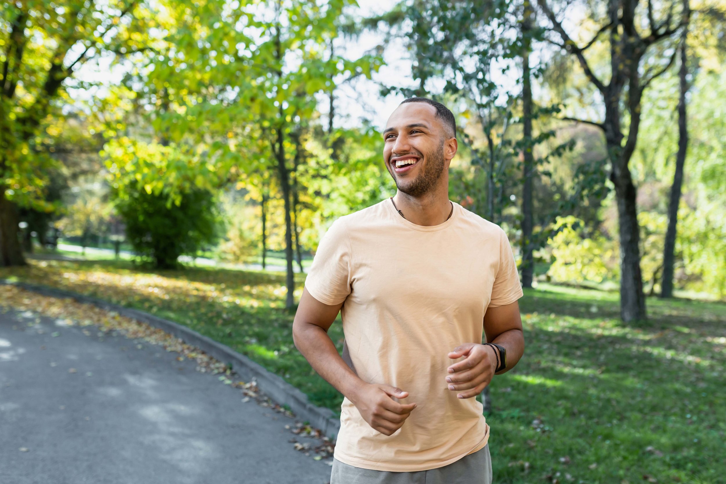 A young man smiling and jogging in a park with green trees and a winding path.
