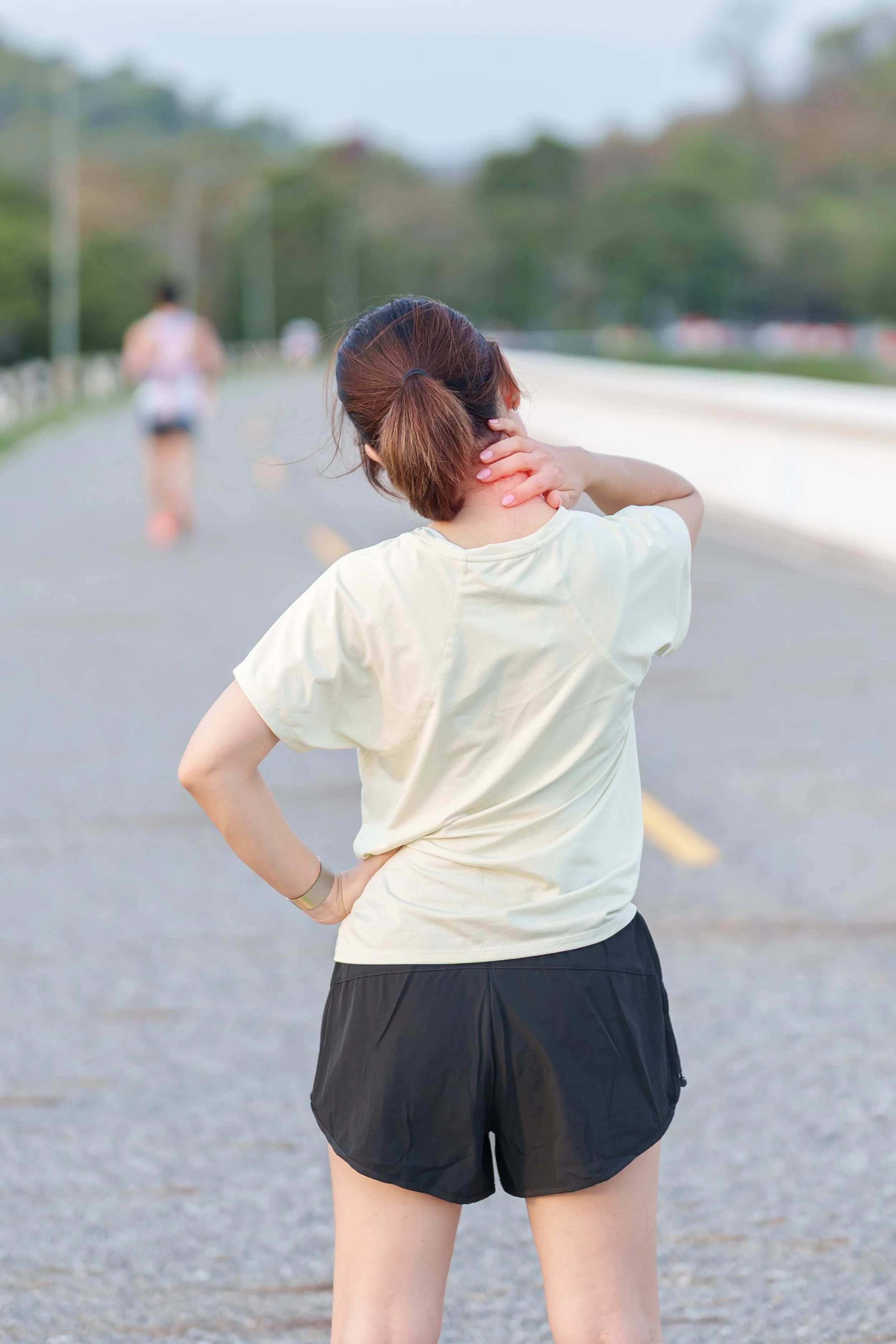 A woman with her hand on her neck, standing on a road and stretching her neck, with another woman jogging in the background.