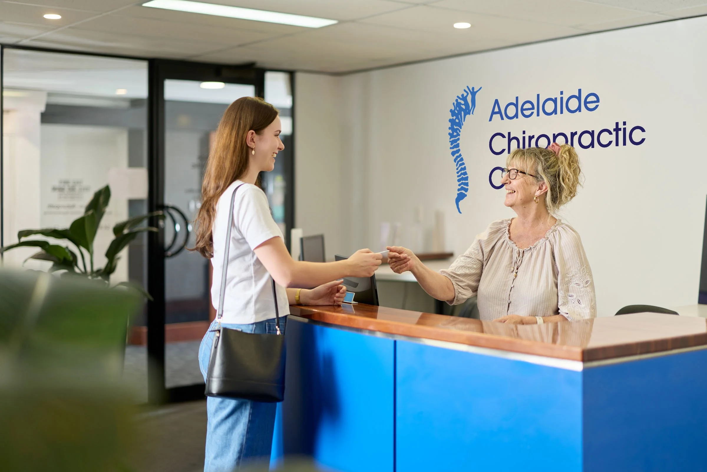 Reception area at Adelaide Chiropractic Clinic where a young woman is smiling and handing a card to an older woman behind the desk, with the clinic's sign and logo on the wall.
