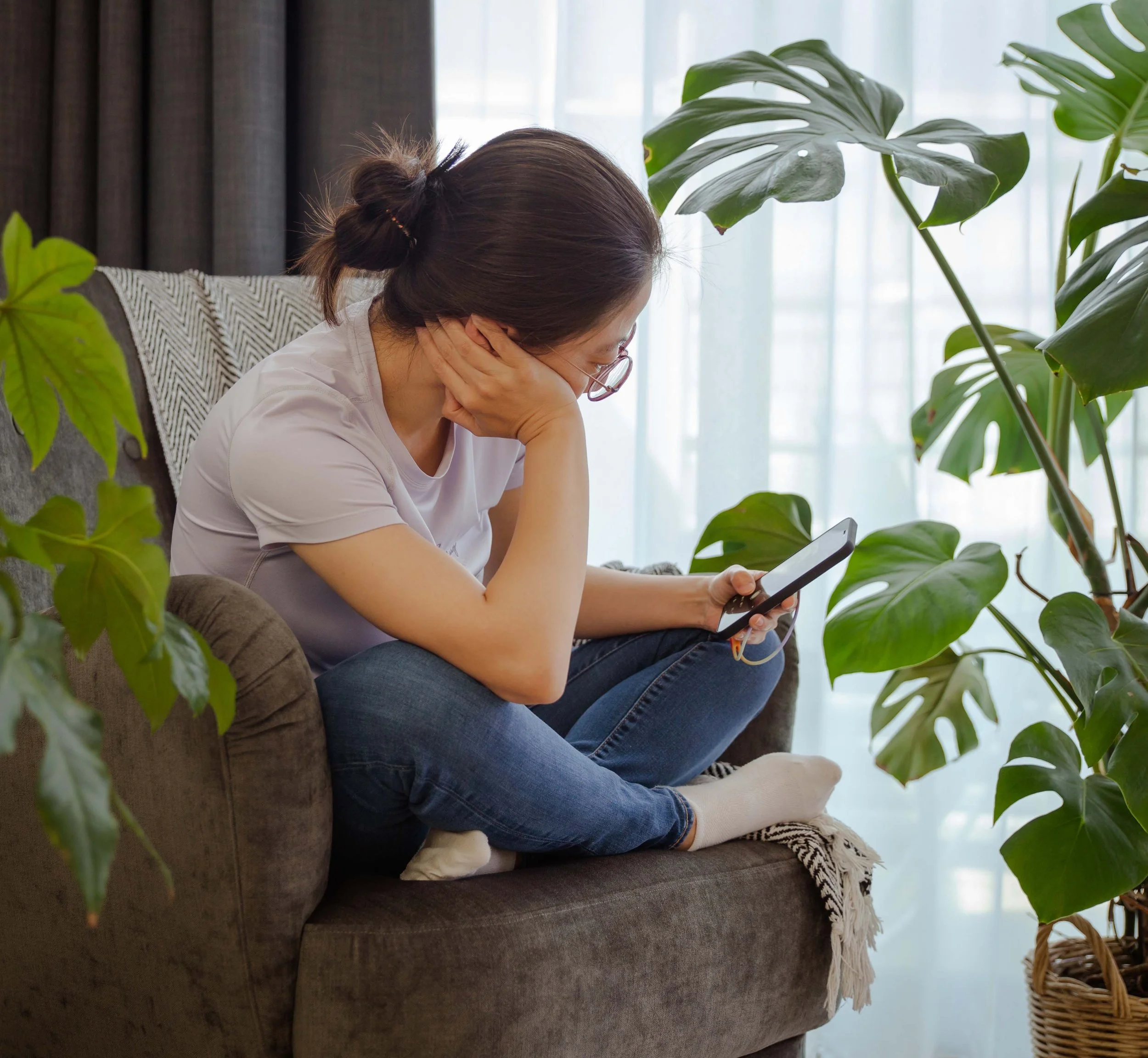 A woman with brown hair, glasses, wearing a white shirt and blue jeans, sitting on a brown sofa with her elbows on her knees, holding her head with one hand while looking at her phone, surrounded by large green houseplants in front of a glass window with sheer white curtains.
