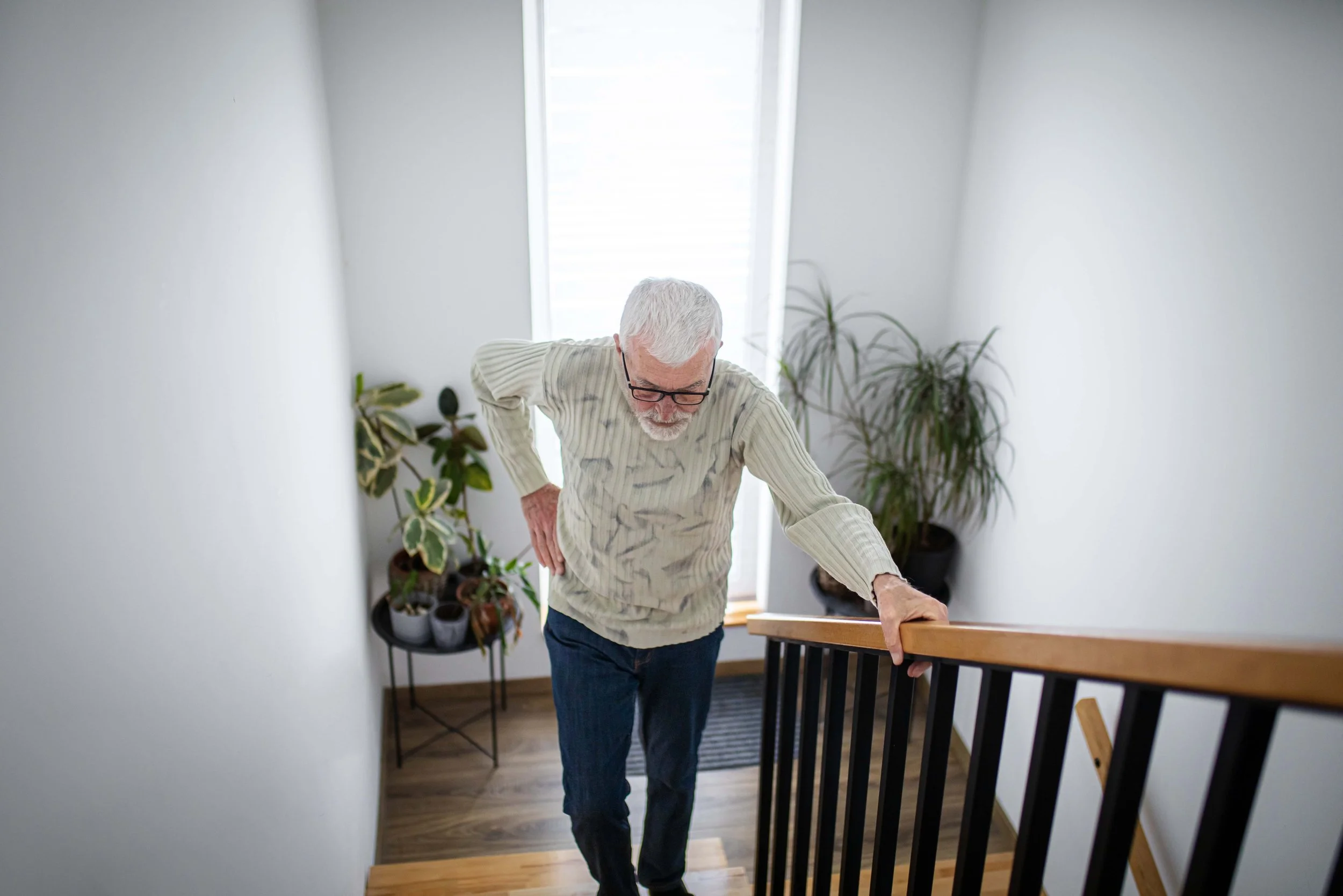 An elderly man with white hair and glasses is ascending a staircase, leaning on the railing. He is wearing a beige striped sweater and dark pants. The staircase is in a well-lit area with white walls and several potted plants in the background.