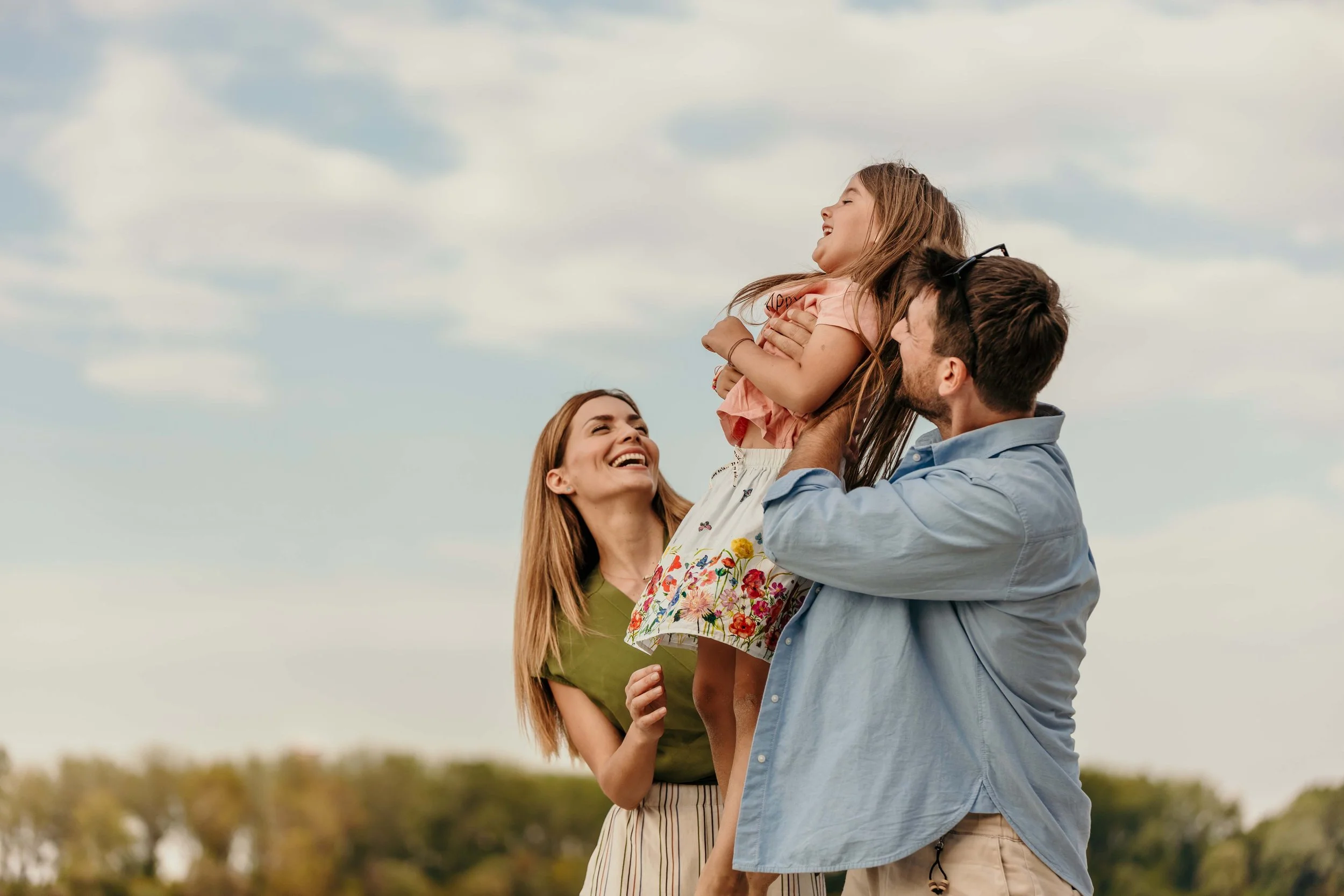 A family enjoying an outdoor moment; a man lifts a girl, both smiling, while a woman looks on joyfully, with a blue sky and trees in the background.