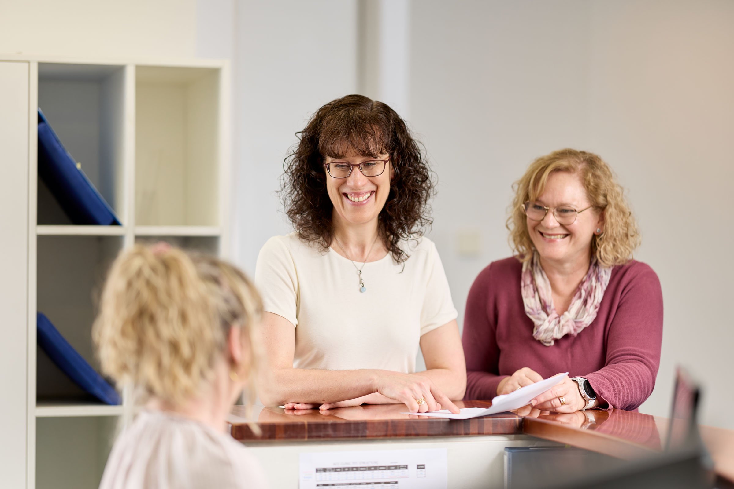 Two women sitting at a table, smiling, engaging in conversation with children in a room with white walls and a bookshelf in the background.