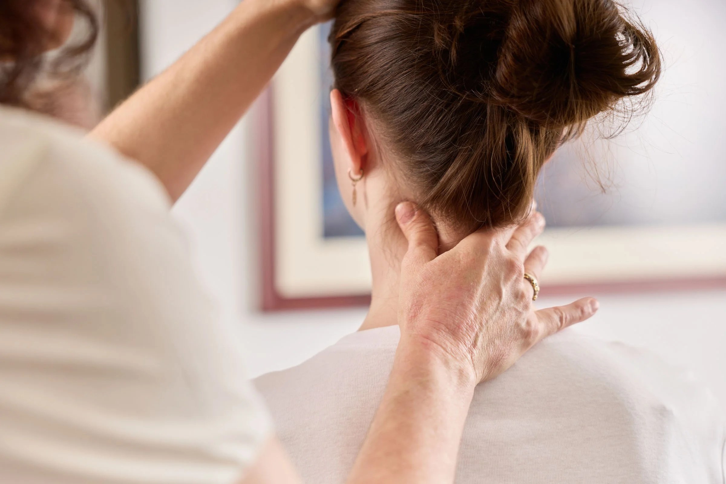 A person with short hair is gently holding the neck of a woman with brown hair styled up, as she tilts her head back slightly. The scene appears to be a neck or head massage or therapy session.
