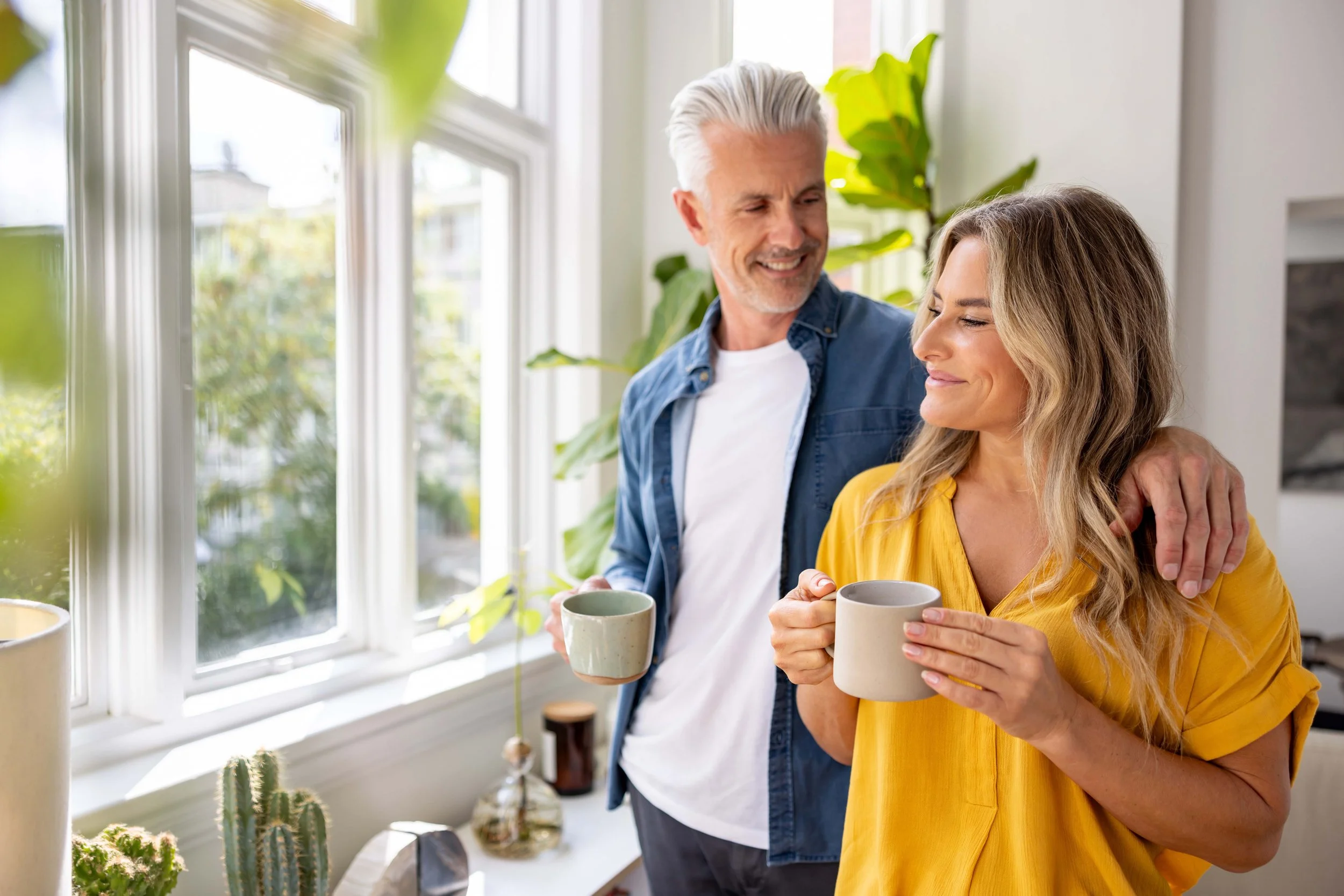 A middle-aged man with gray hair and a woman with long wavy hair hold coffee mugs in a kitchen with large windows and green plants, smiling and enjoying the moment.