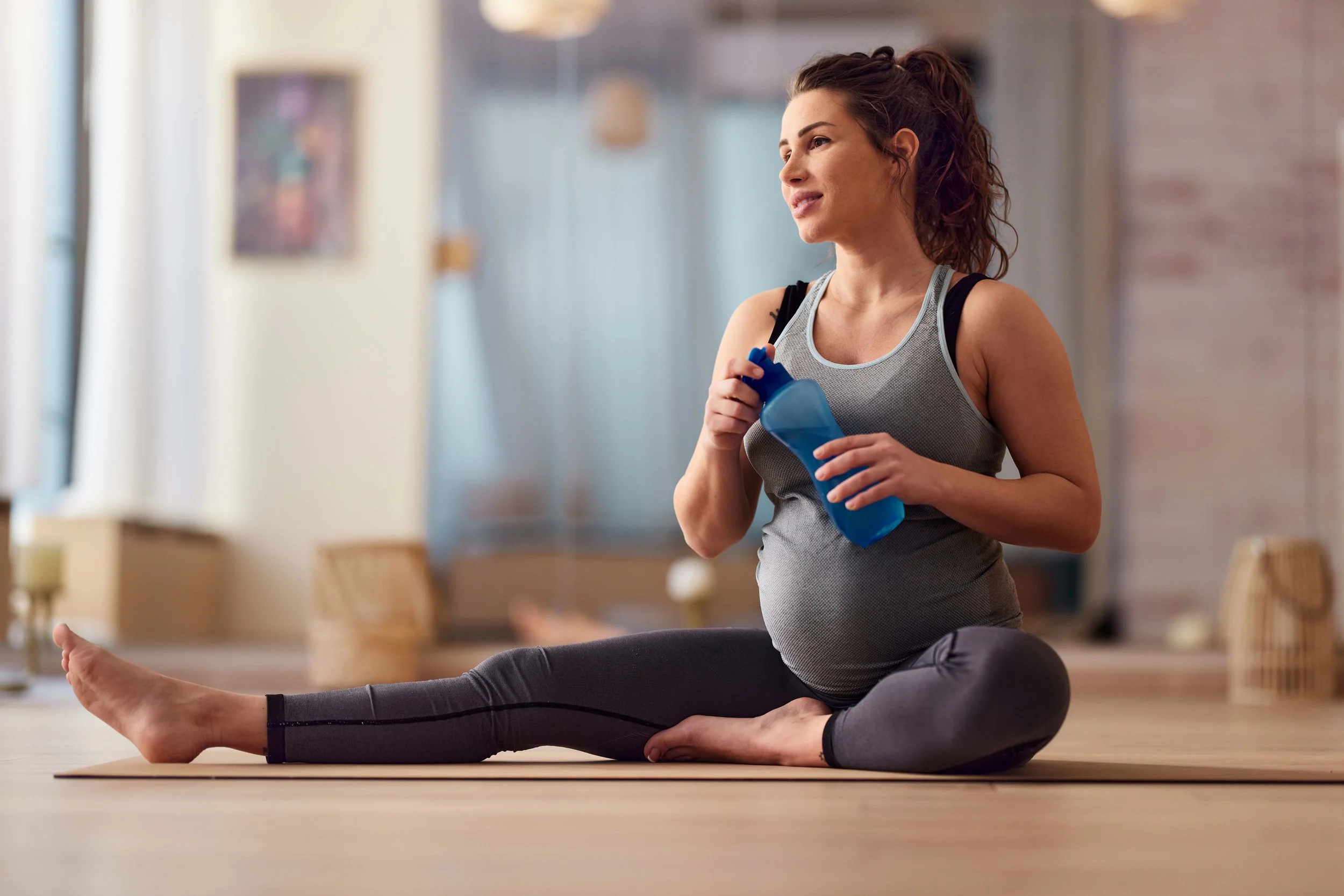 A woman performing yoga indoors, sitting in a cross-legged position with her arms relaxed, holding a water bottle, and wearing athletic clothing.