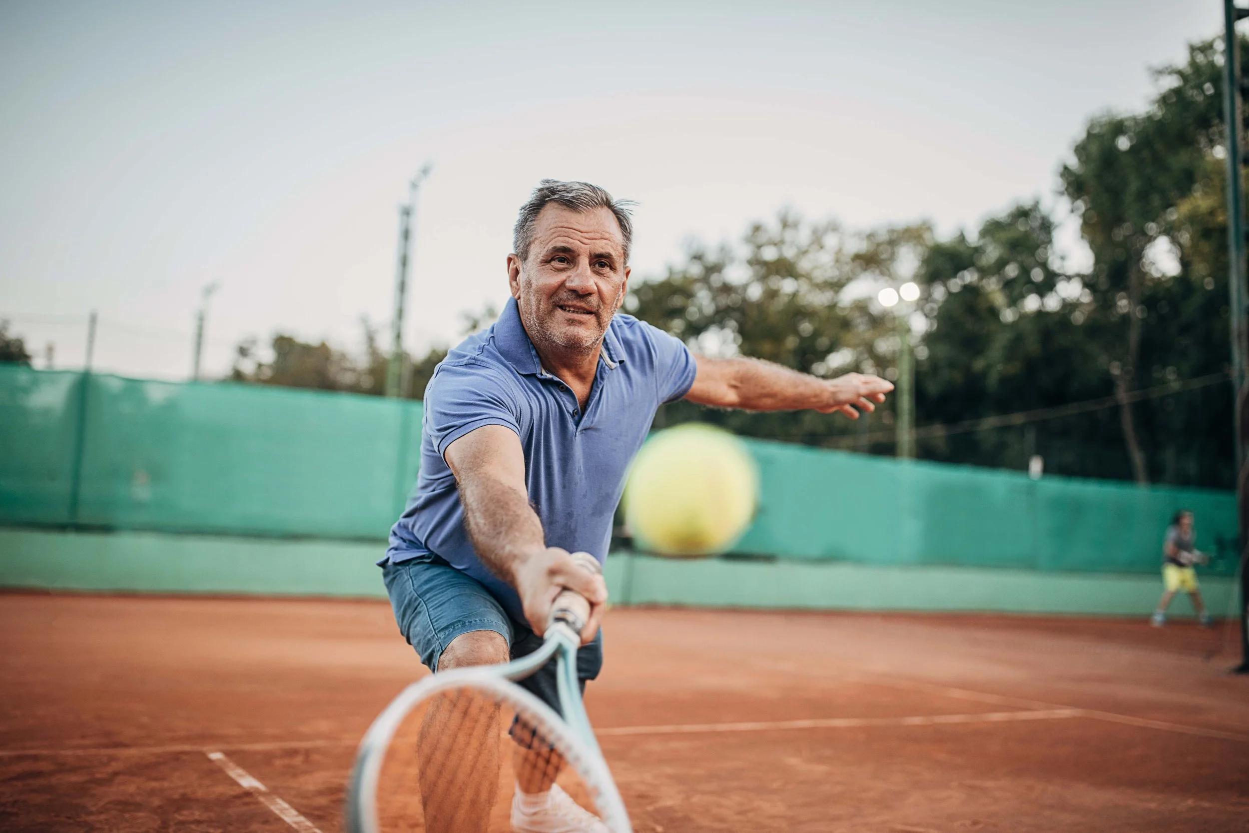 An older man playing tennis on a clay court during daytime, reaching to hit a ball with a racquet, wearing a blue polo shirt and shorts, with trees and a green windscreen in the background.