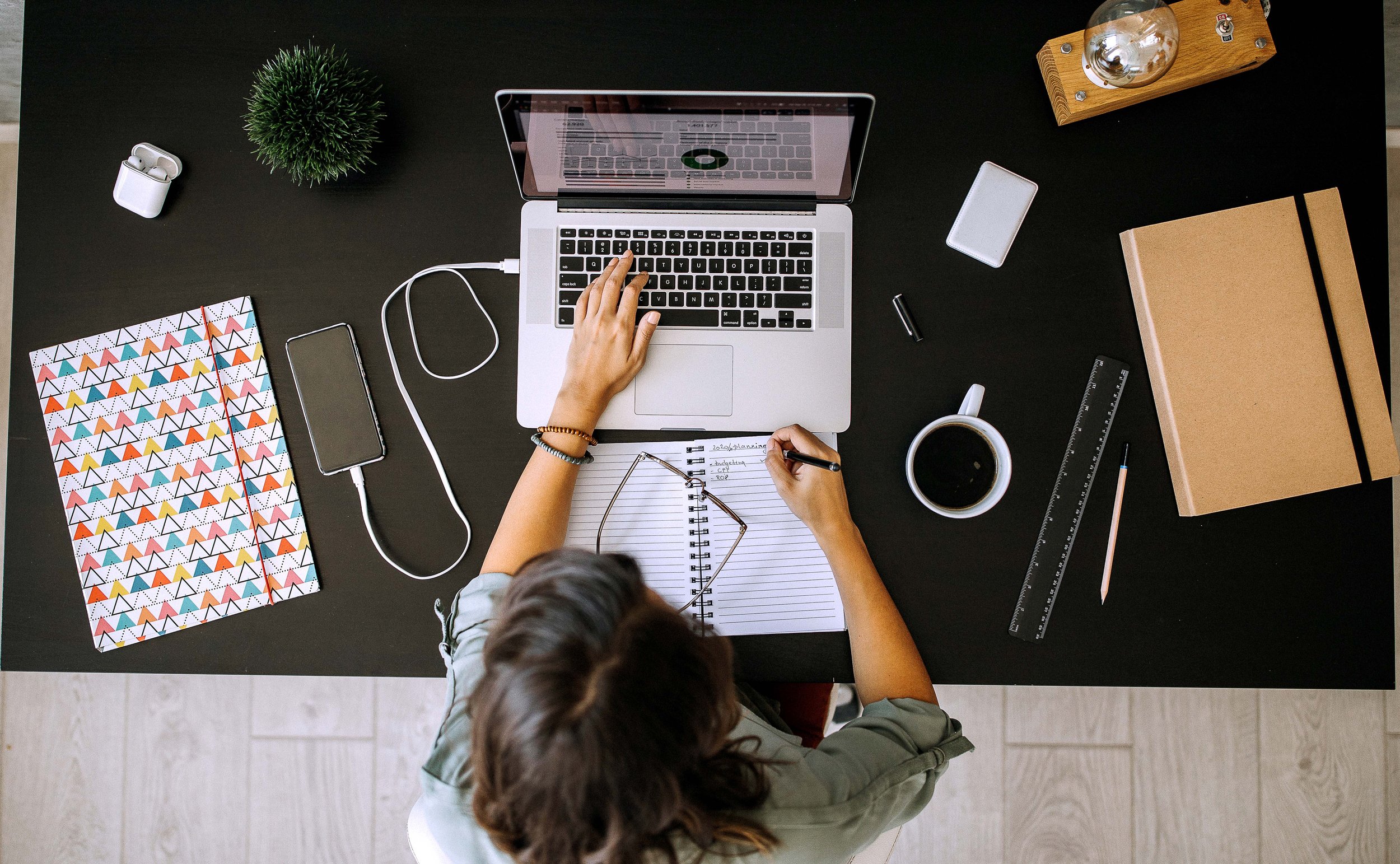 Overhead view of a person working at a desk with a laptop, a notebook, glasses, a coffee cup, a ruler, a pen, a notebook with a colorful geometric pattern, a small plant, a light bulb, and a white wallet.