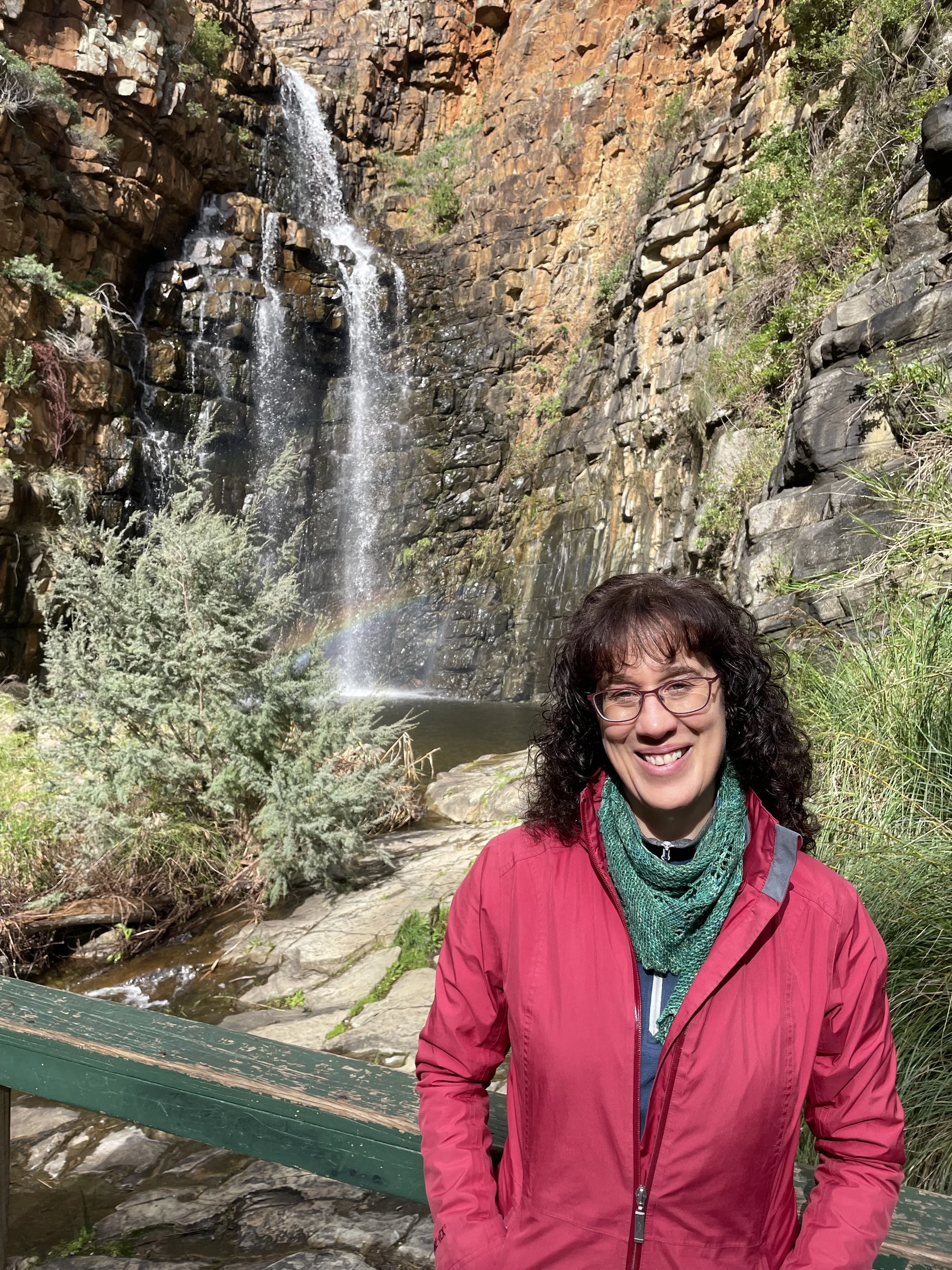 A woman with glasses and curly dark hair smiling in front of a waterfall cascading down a rocky cliff with sparse greenery, wearing a red jacket and green scarf, near a small stream.