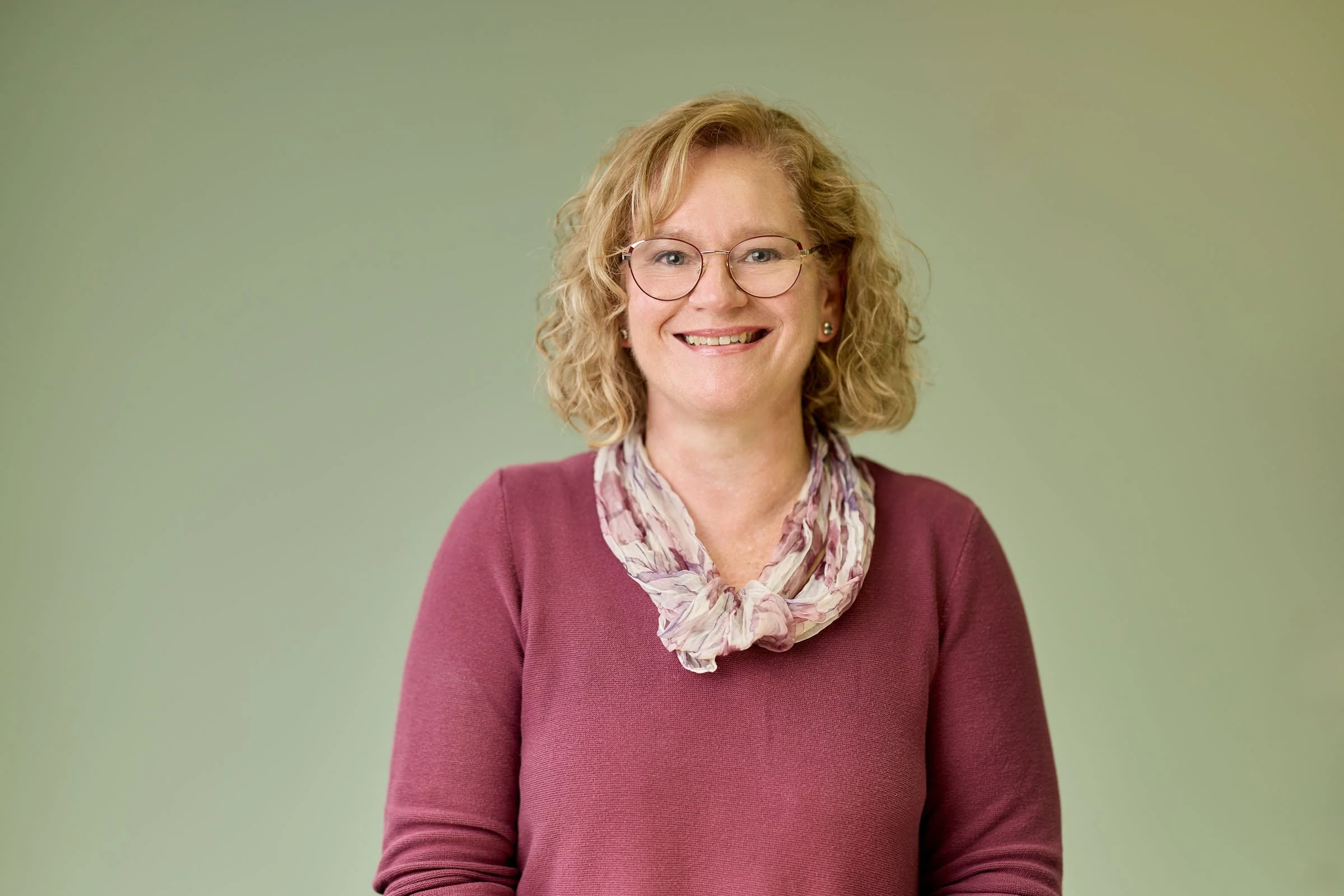 A middle-aged woman with curly blonde hair, wearing glasses, a pinkish-purple sweater, and a colorful scarf, smiling against a plain green background.