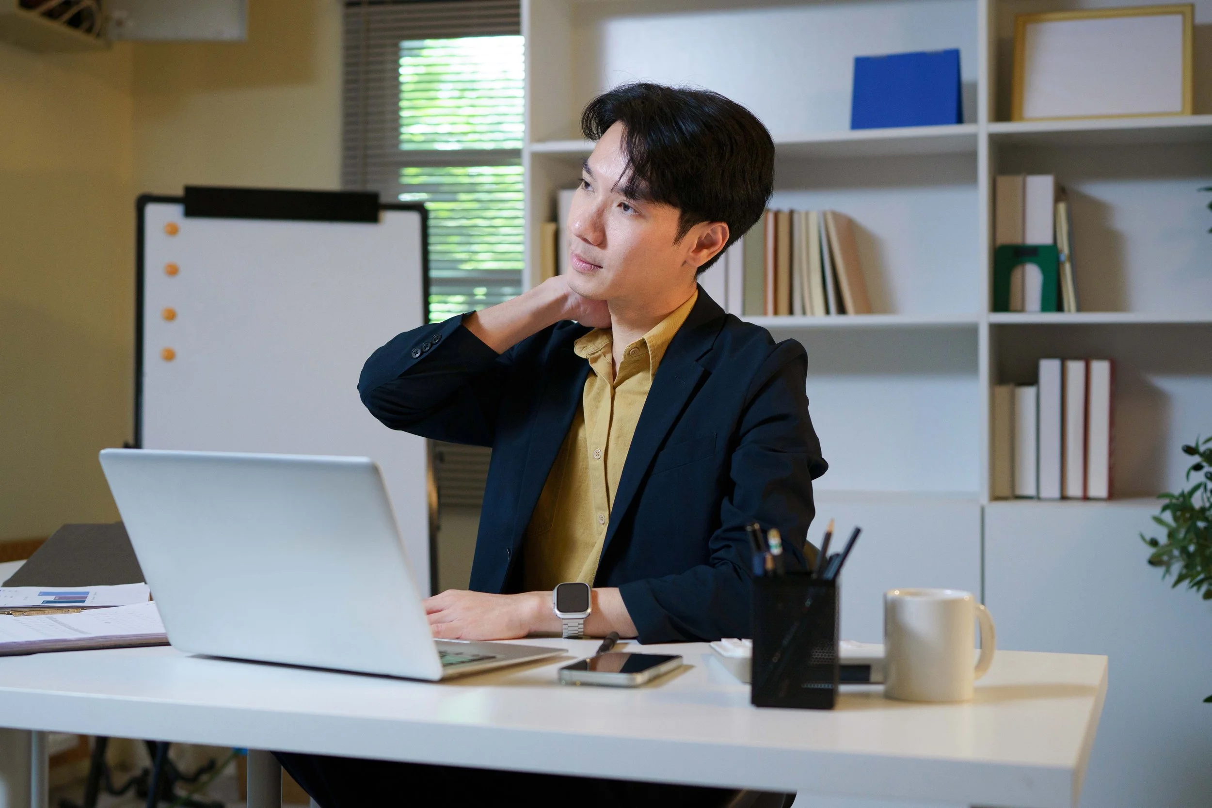 A man in business attire sits at a desk with a laptop, contemplating, with bookshelves and a window in the background.