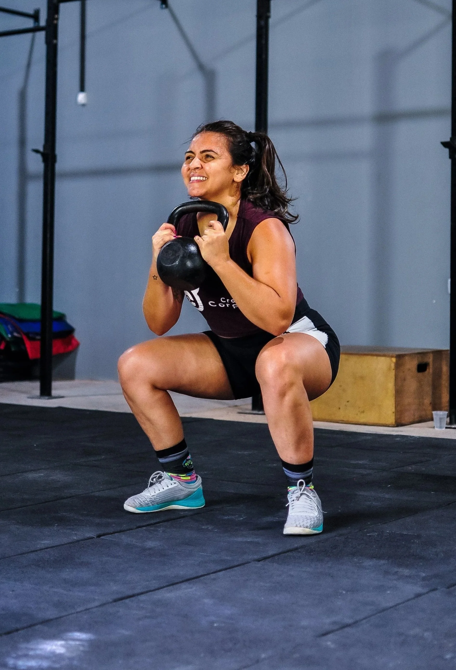 A woman lifting a kettlebell during a workout at a gym, squatting with a smile on her face.