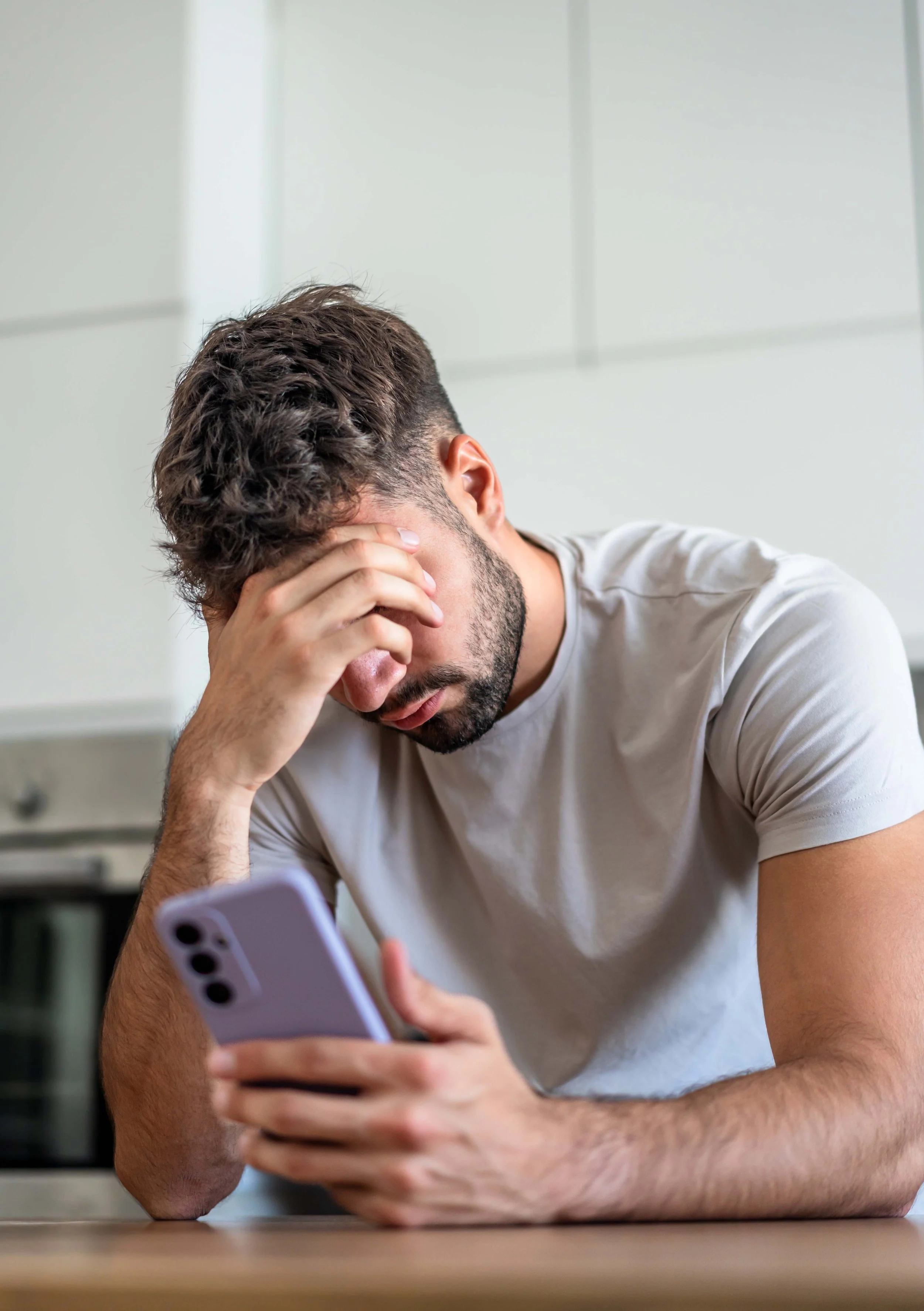 A young man with brown hair and a beard is sitting at a table, holding his head with his left hand, and looking at his phone. He appears distressed or frustrated, with his eyes closed and his facial expression tense.