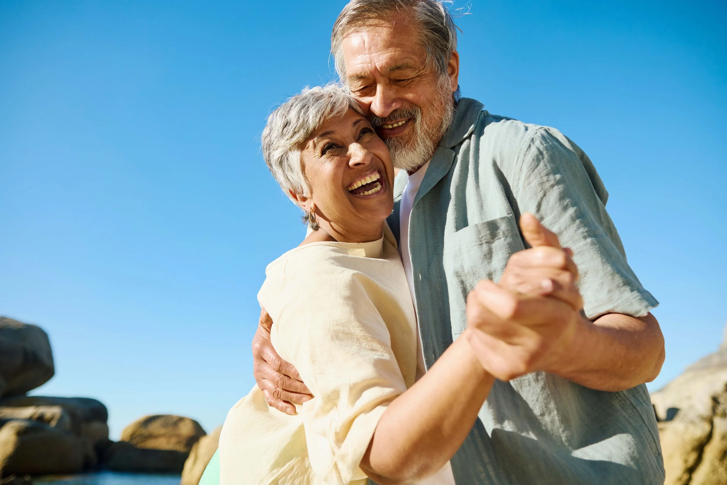 An elderly couple smiling and hugging outdoors against a backdrop of bright blue sky and rocky terrain.