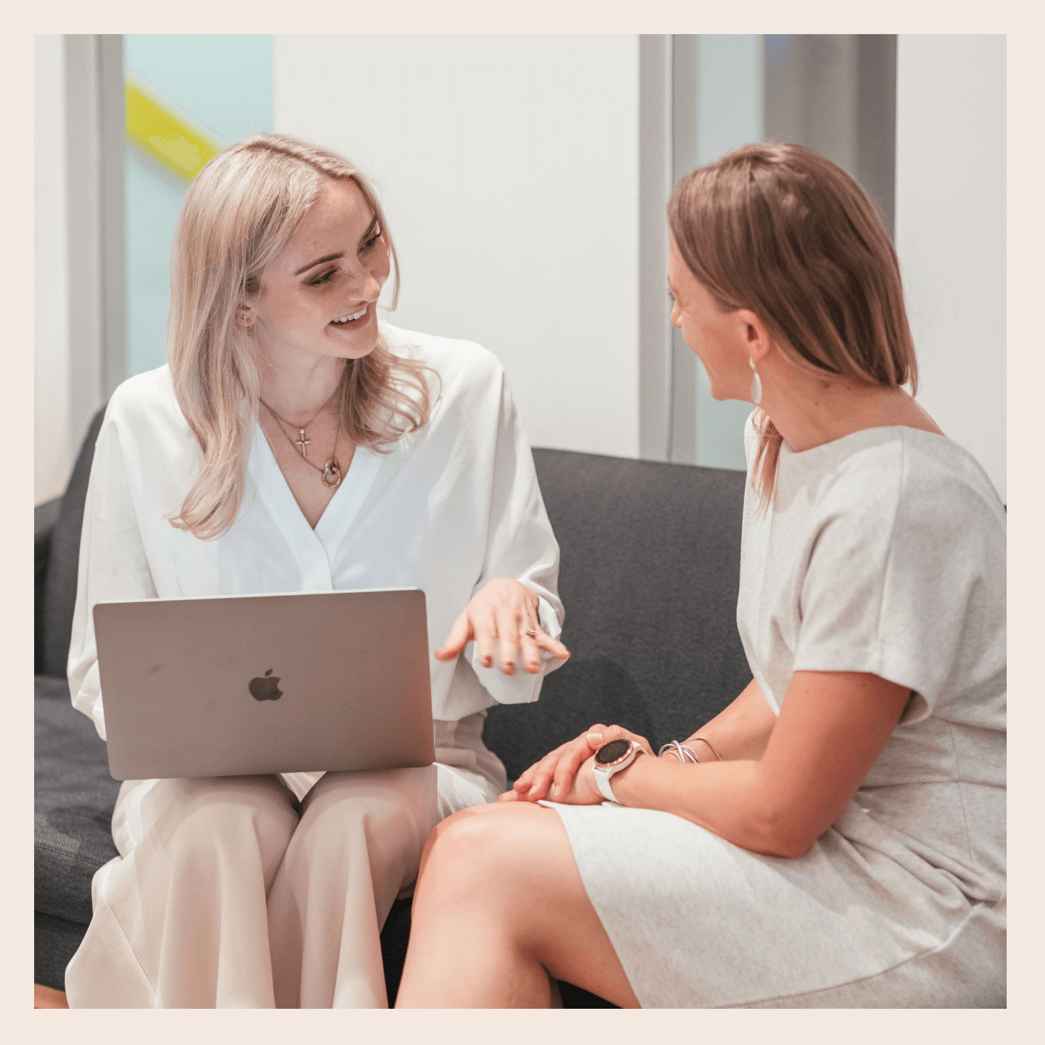 Two women are sitting on a gray couch in an office, engaging in a conversation. One woman is smiling and gesturing with her hand, while the other woman is listening, wearing a watch and earrings. The woman on the left has blonde hair and is holding a silver laptop, while the woman on the right has brown hair and is wearing a light-colored dress.