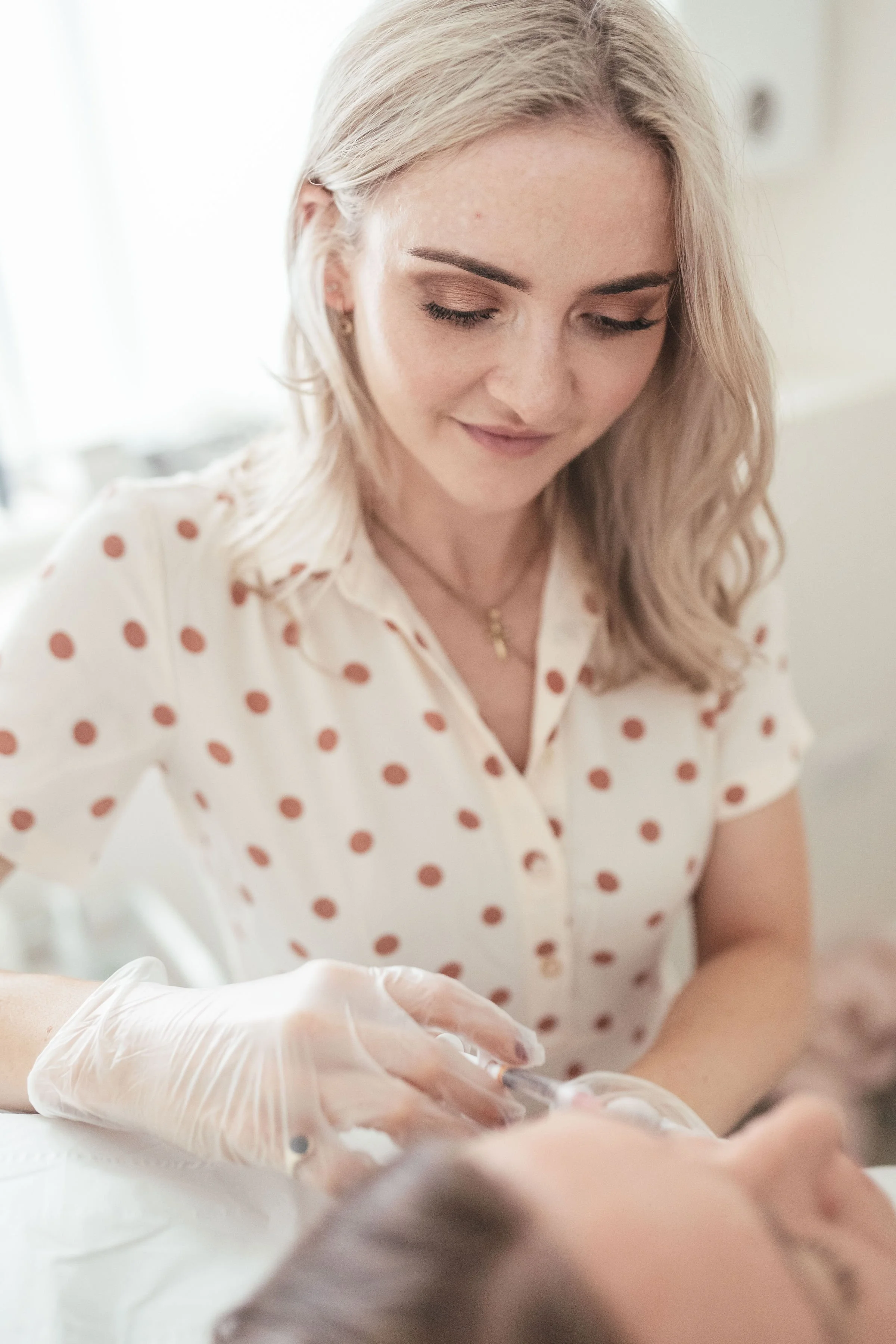 A woman with blonde hair, wearing a white shirt with brown polka dots, is looking down at a person lying on a surface, with medical gloves on her hands, administering an injectable treatment.