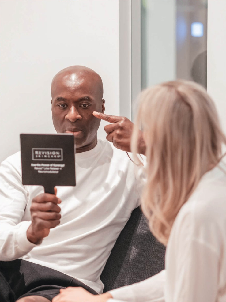 A man holding a mirror and pointing at his face while talking to a woman with blonde hair in a white shirt, in a medical or consultation setting.