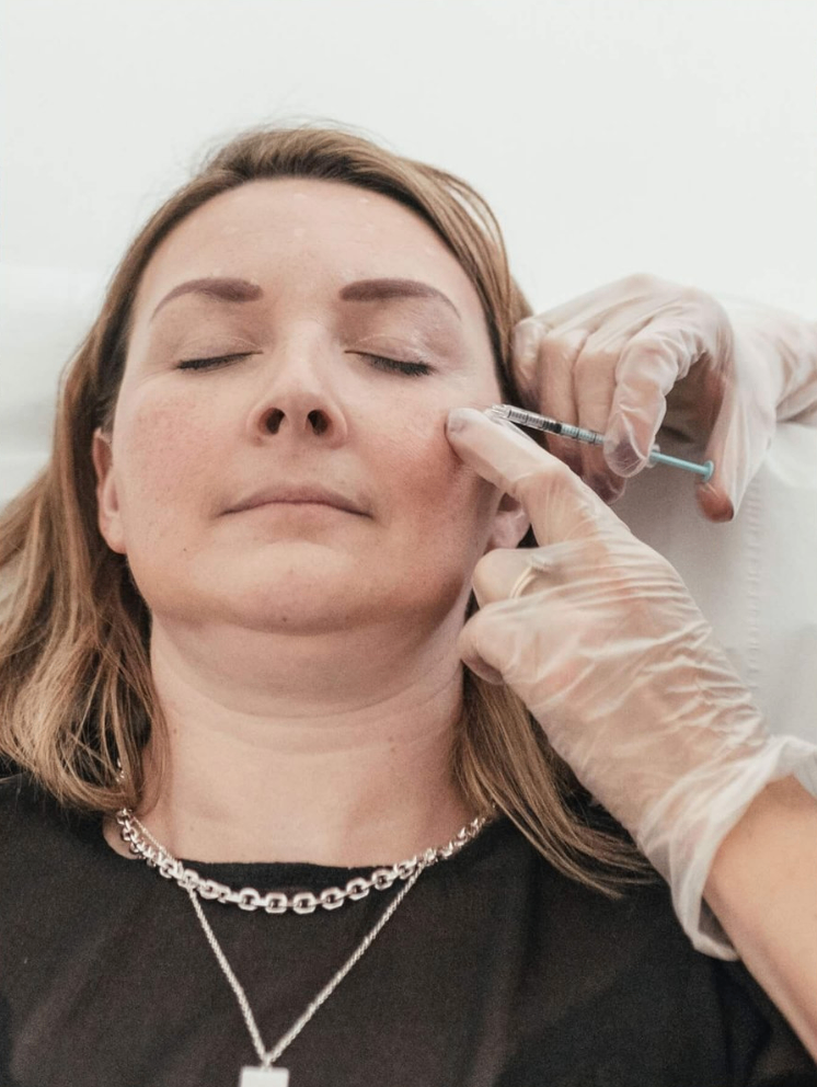 A woman with closed eyes receiving a facial injection from a medical professional wearing gloves.