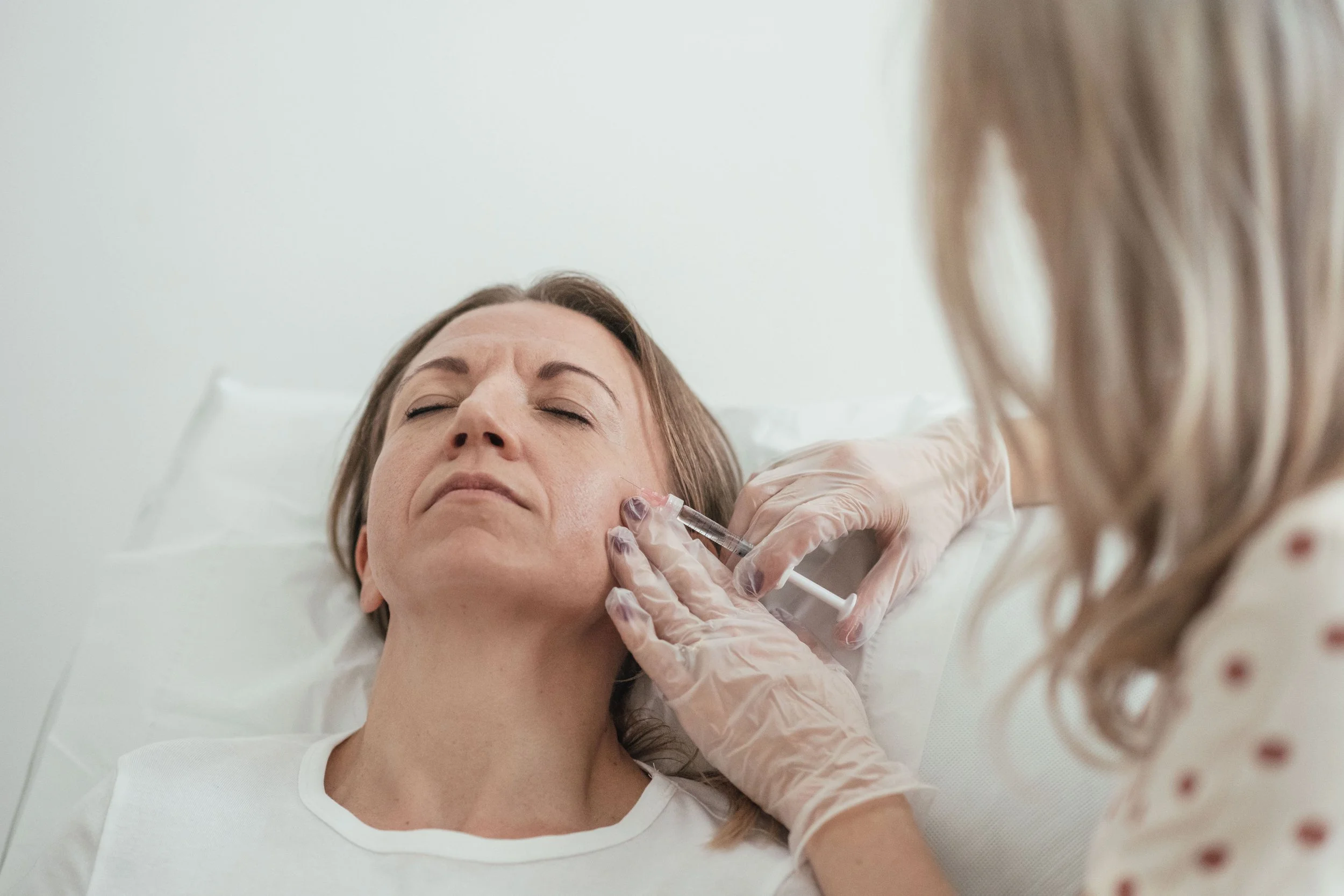 A woman lying in a hospital bed receiving a facial injection from a medical professional wearing gloves, with her eyes closed.