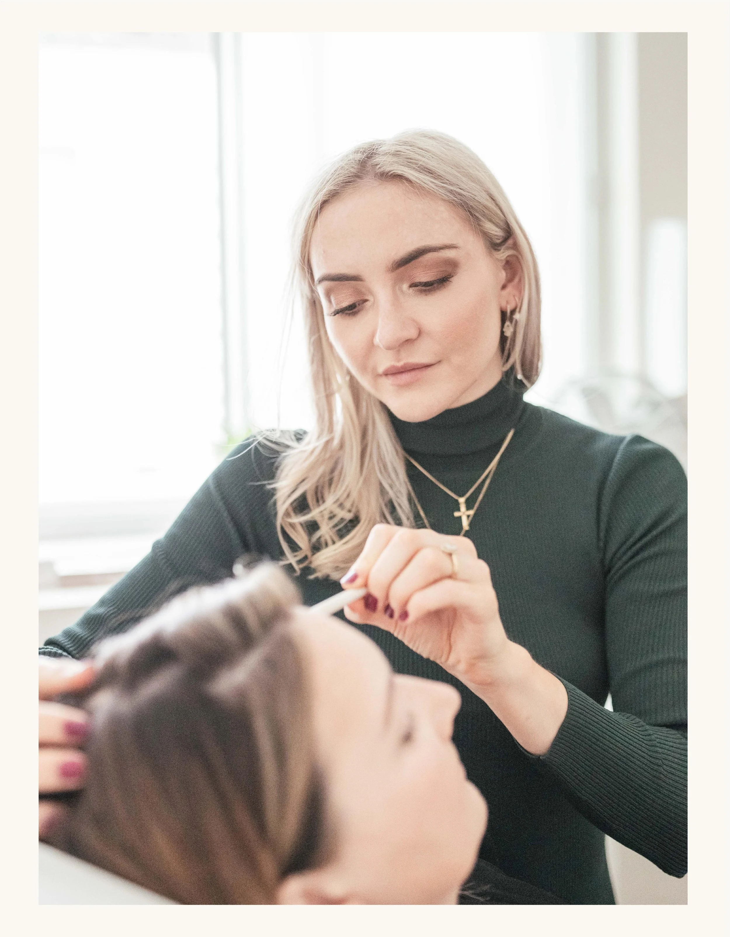 Dr Catharine Denning wearing a black top and gold jewelry, including a cross necklace and star earrings, performing a skin rejuvenation treatment in a bright room with large windows.