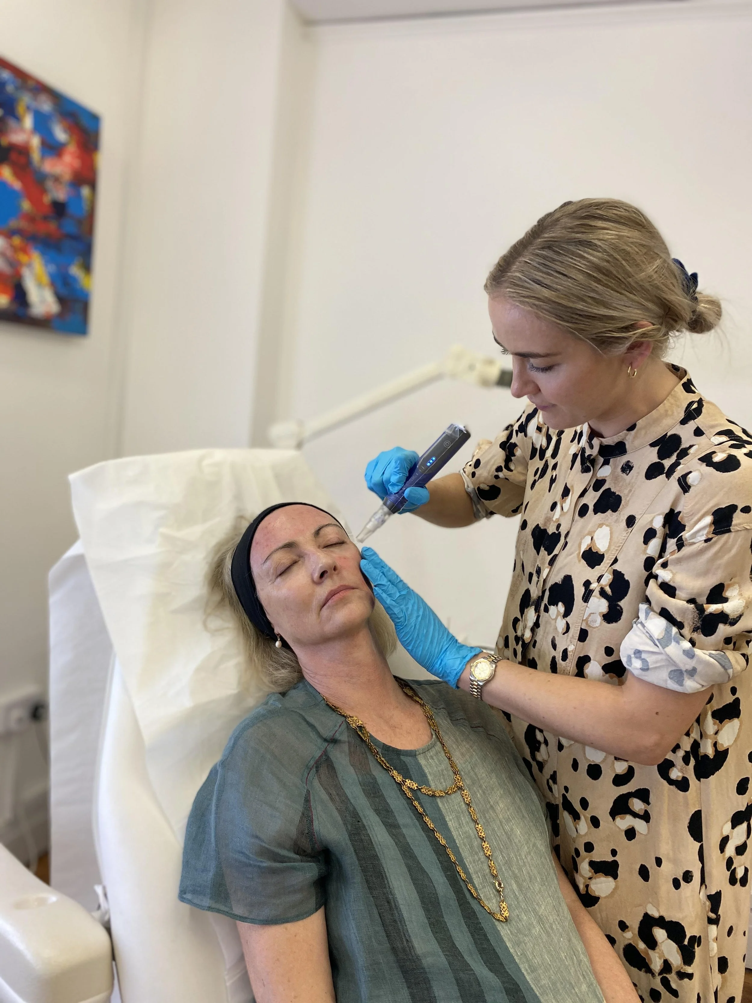 A woman with blonde hair lying on a hospital bed, receiving a cosmetic treatment from a female medical professional in a leopard print dress. The medical professional is using a syringe or similar device, wearing blue gloves, and the patient looks relaxed with her eyes closed.