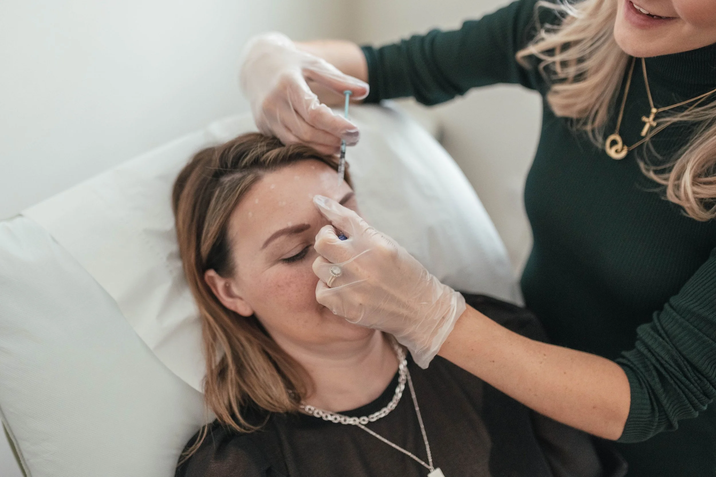 A woman lying on a medical bed receiving a cosmetic injection in her forehead from a healthcare professional wearing gloves.