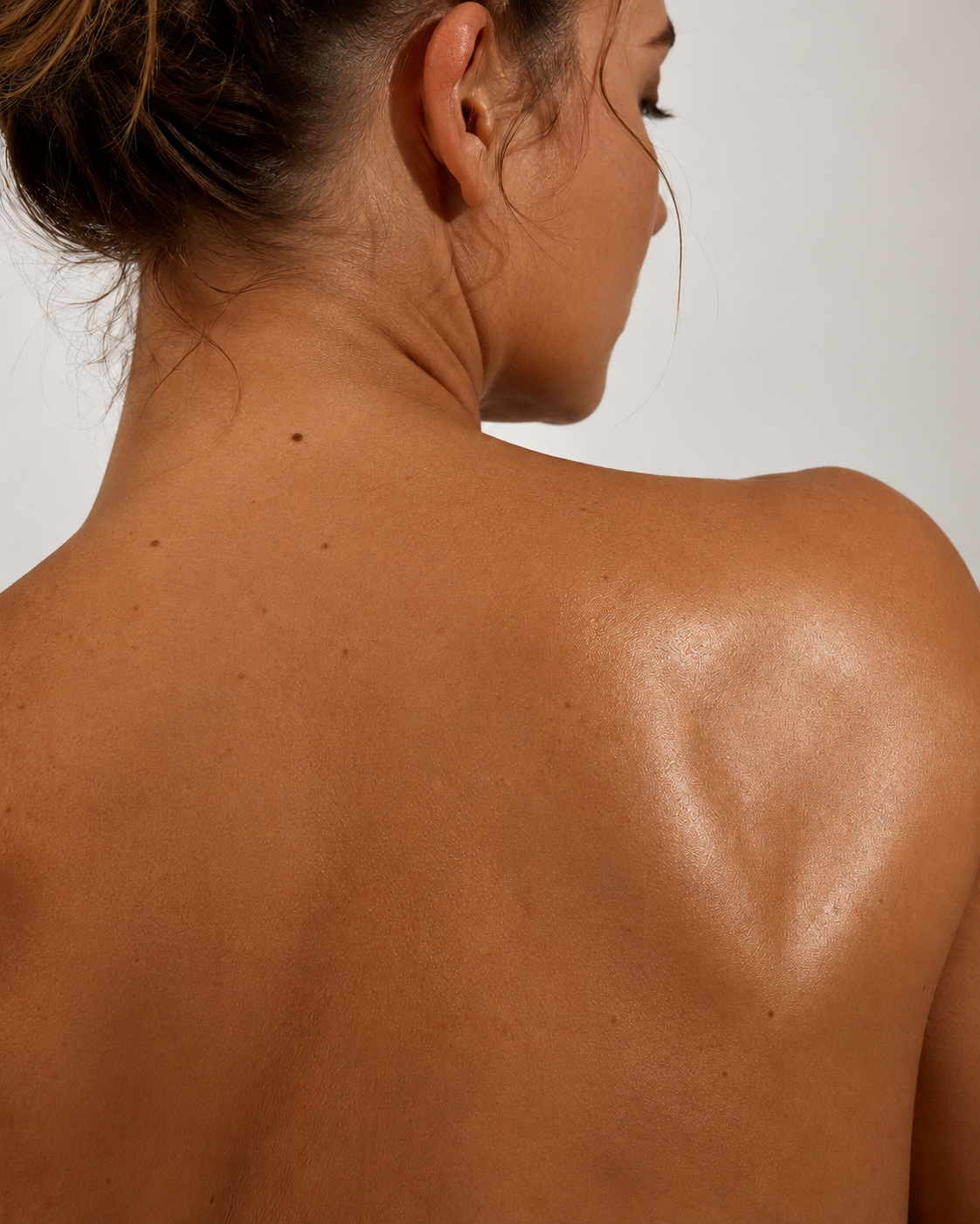 Back view of a woman with tan skin and brown hair tied back, showing her shoulder and neck, with visible mole and small freckles.