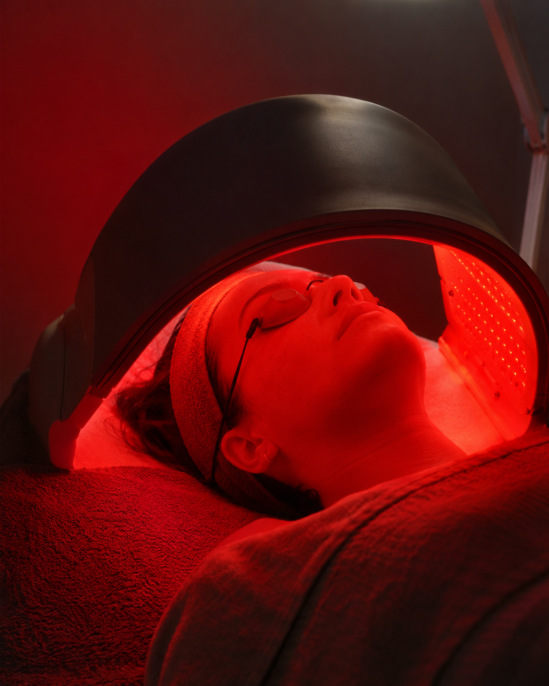 Woman lying on a treatment bed under red light therapy, with a facial mask and eye protection.