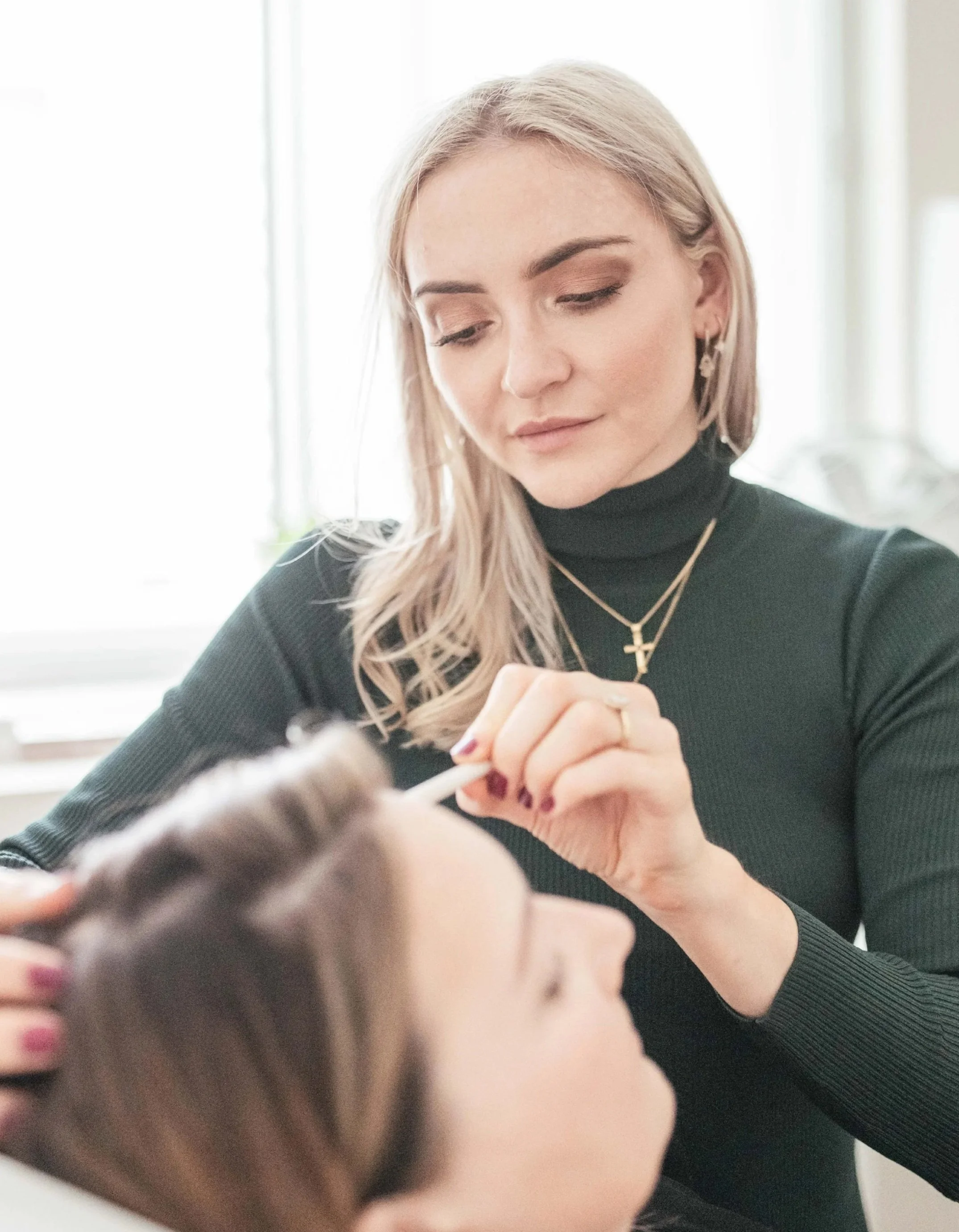 A woman with blonde hair, wearing a black turtleneck, jewelry including cross and floral earrings, is performing an injectable treatment in a clinical setting on a patient.
