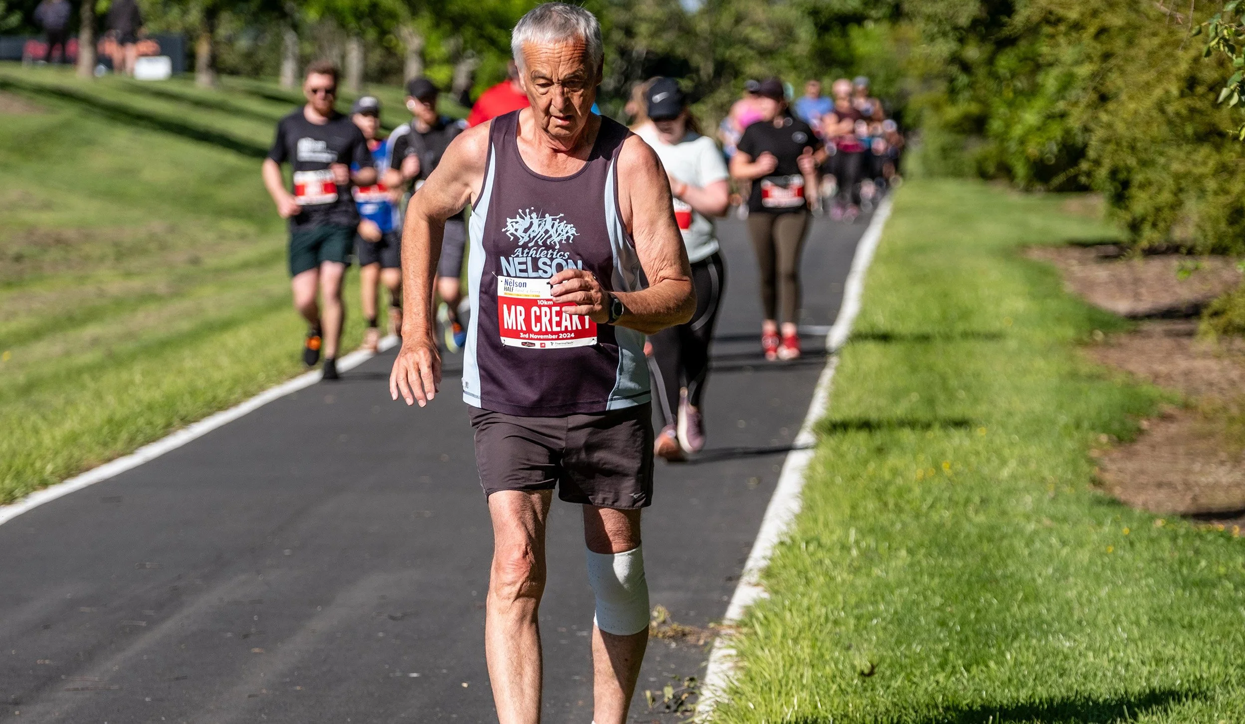 Older man running in a marathon on a paved trail surrounded by green grass and trees, wearing a black athletic tank top and shorts, with a race bib labeled 'MR Creak'.
