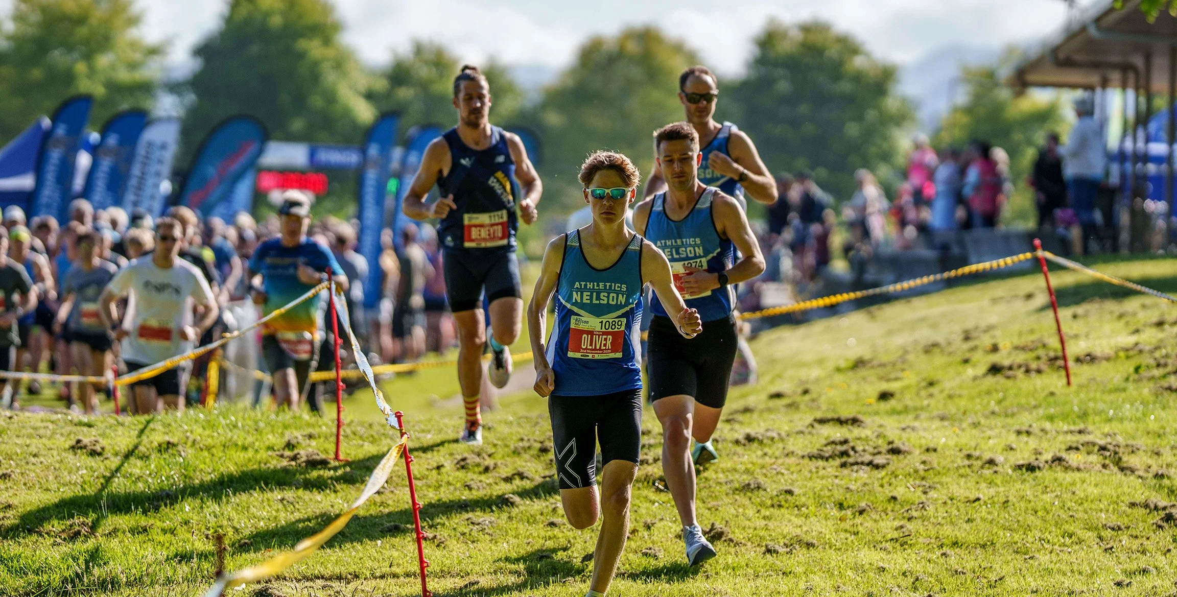 A group of runners participating in a race outdoors, with some wearing athletic gear and sunglasses, on a grassy trail with a crowd of spectators and blue flags in the background.
