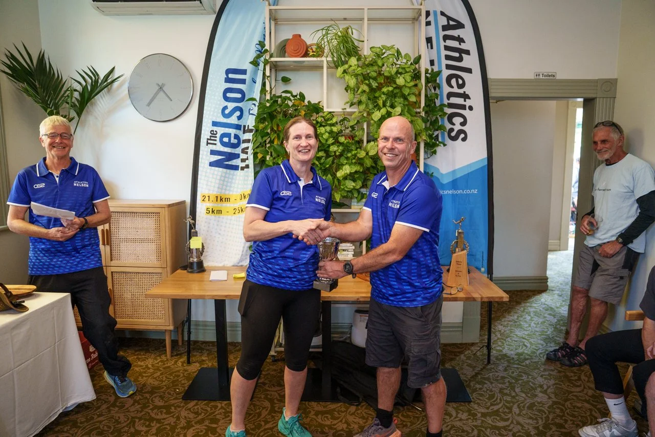Two people in blue shirts shaking hands and holding a trophy at an indoor award ceremony, with a banner, a clock, and people watching.