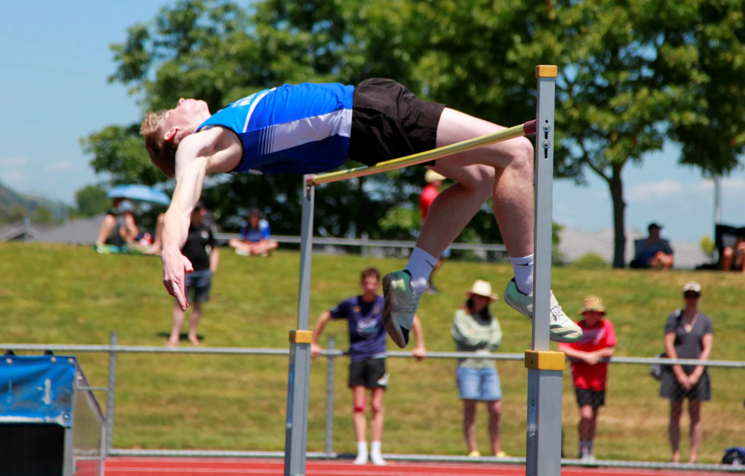 A male athlete in a blue tank top and black shorts clearing a high jump bar outdoors during a track and field event, with spectators and trees in the background.