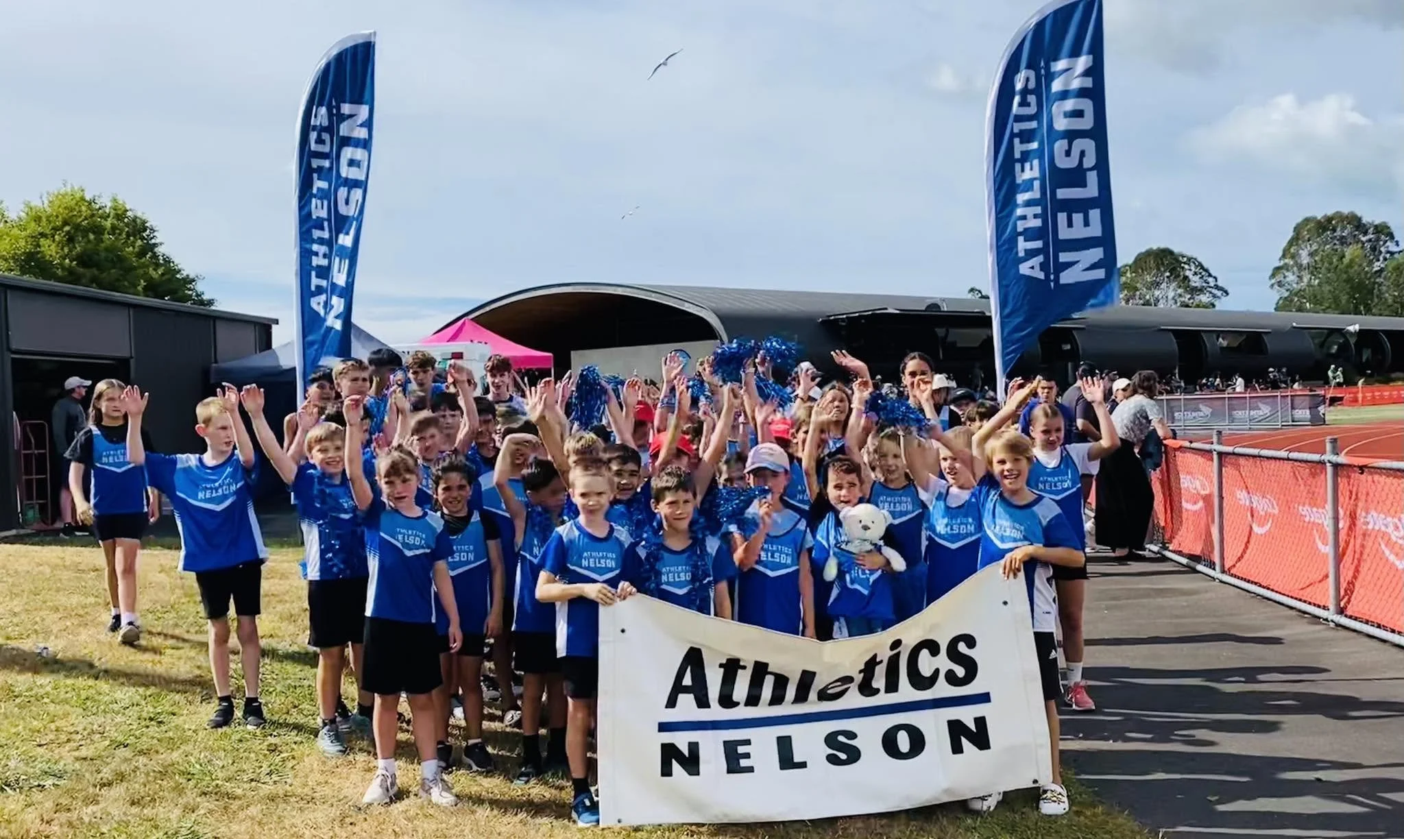 Young athletes in blue uniforms and cheerleading attire gather behind a banner that reads 'Athletics Nelson' at an outdoor sports event. They are on a track field with flags and tents in the background, with some raising their hands cheerfully.
