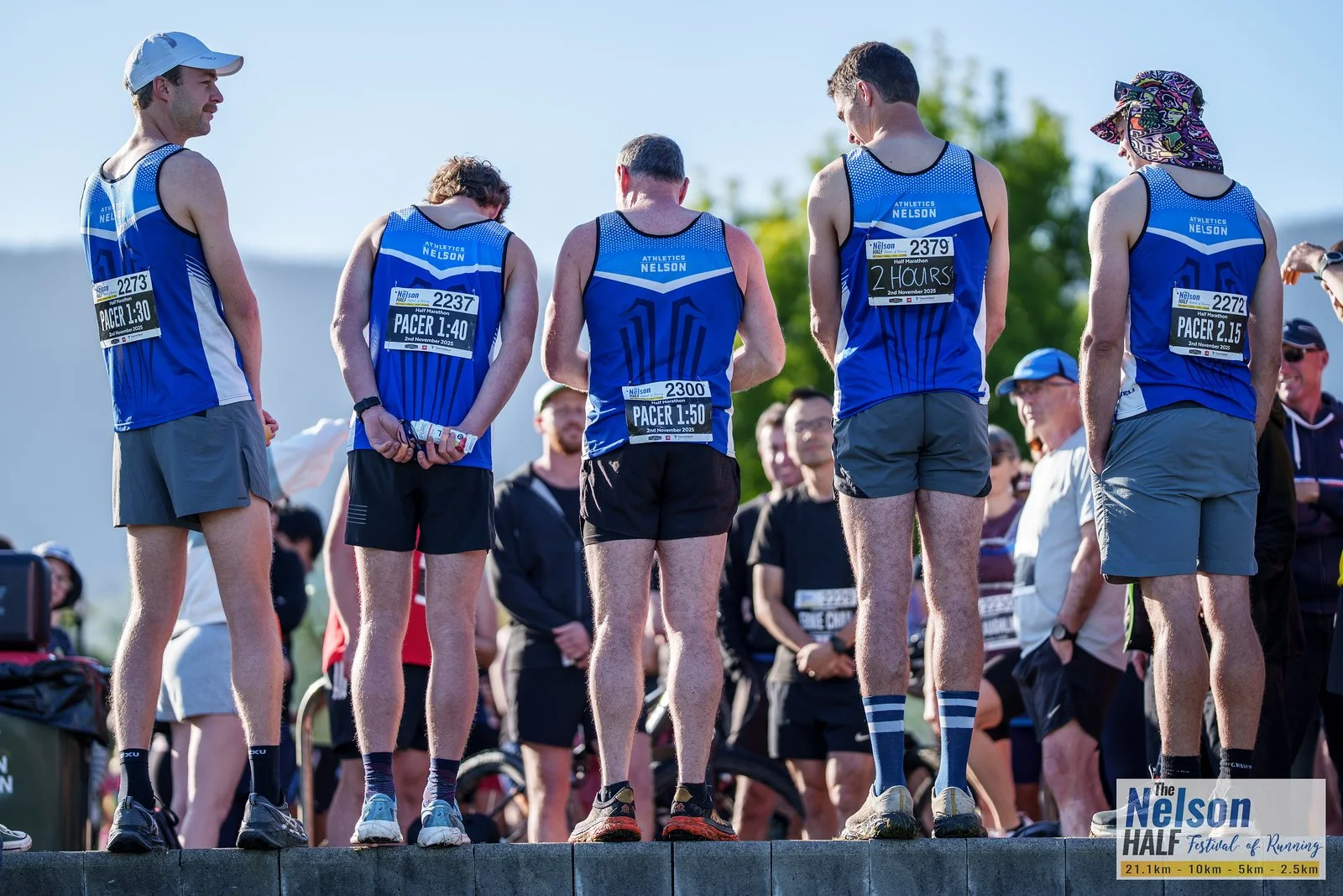 A group of five runners wearing blue athletic shirts with race numbers, gathered at the start of a race, with spectators in the background at an outdoor event during daytime.