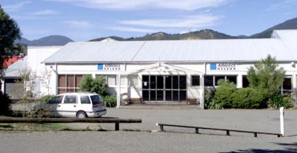 A white industrial or commercial building with a peaked roof, surrounded by bushes and a parking lot with an SUV parked in front. Mountains are visible in the background.