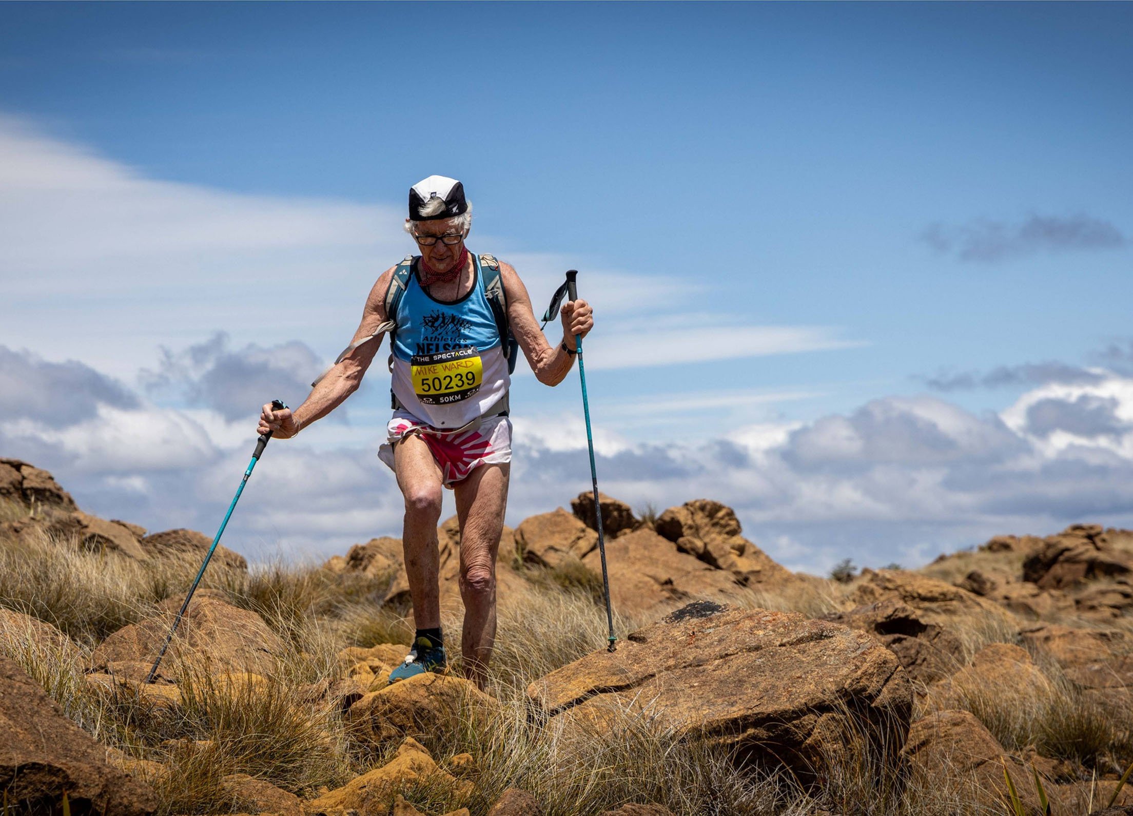 An elderly man wearing a blue running tank top with a race bib and helmet, using trekking poles while hiking on rocky terrain with grass, under a partly cloudy sky.