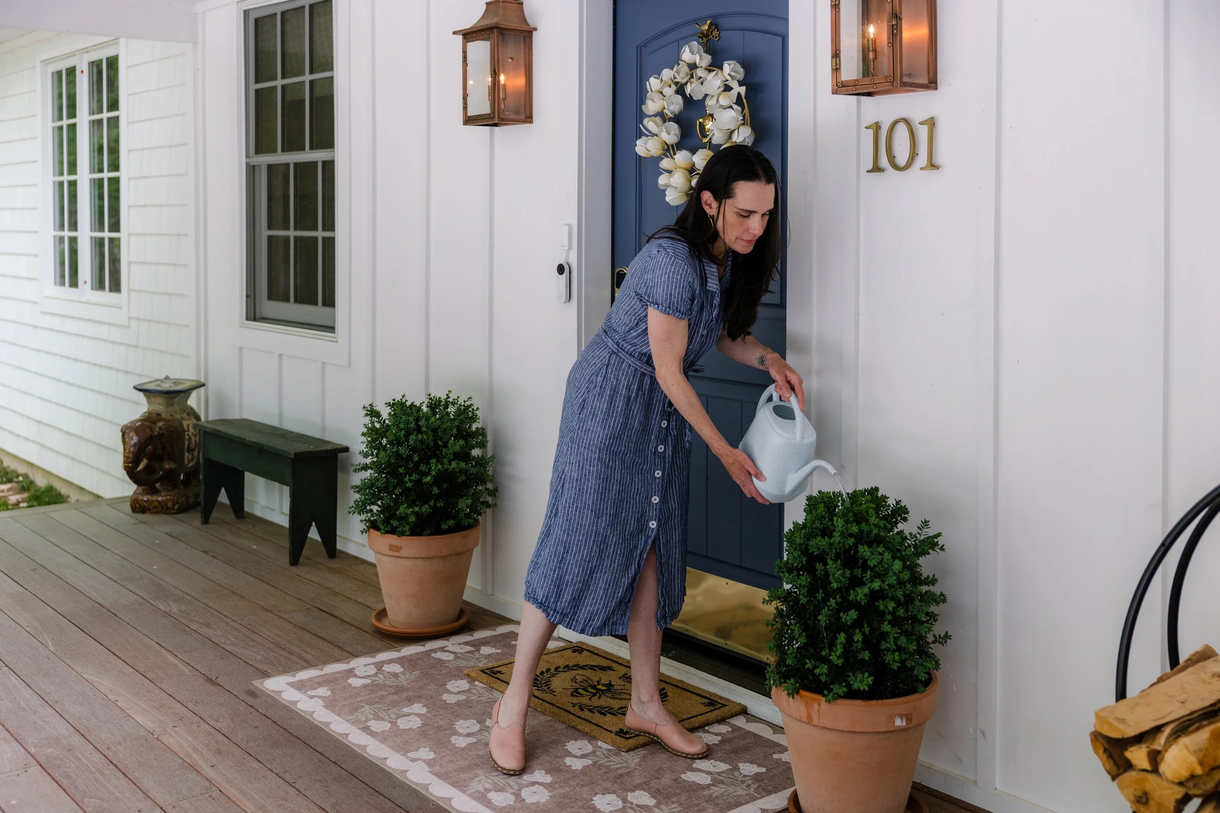 A woman watering plants on her front porch, standing on a black and yellow doormat in front of a blue door with a white floral wreath. The porch has a beige and white patterned rug, potted plants, and copper wall lanterns.