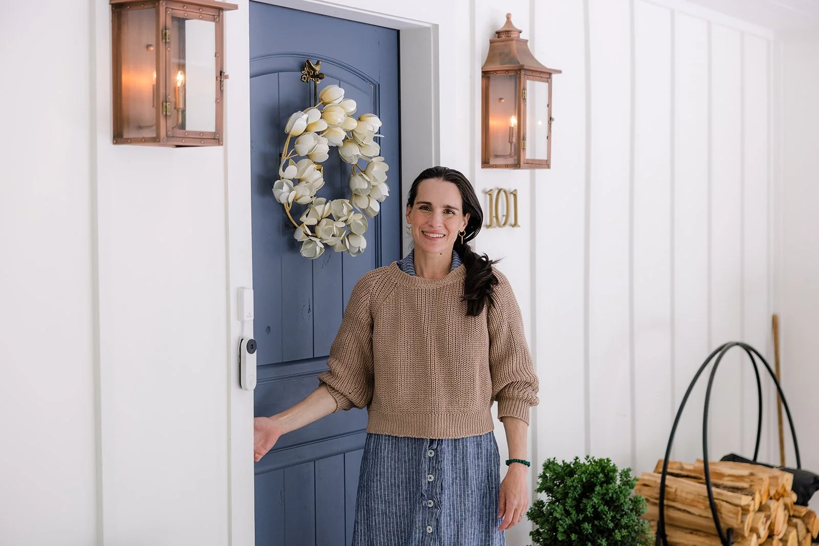 A woman with dark hair and a smile, standing at a blue front door decorated with a white floral wreath, next to a wall with the house number 101 and copper-colored outdoor lanterns.