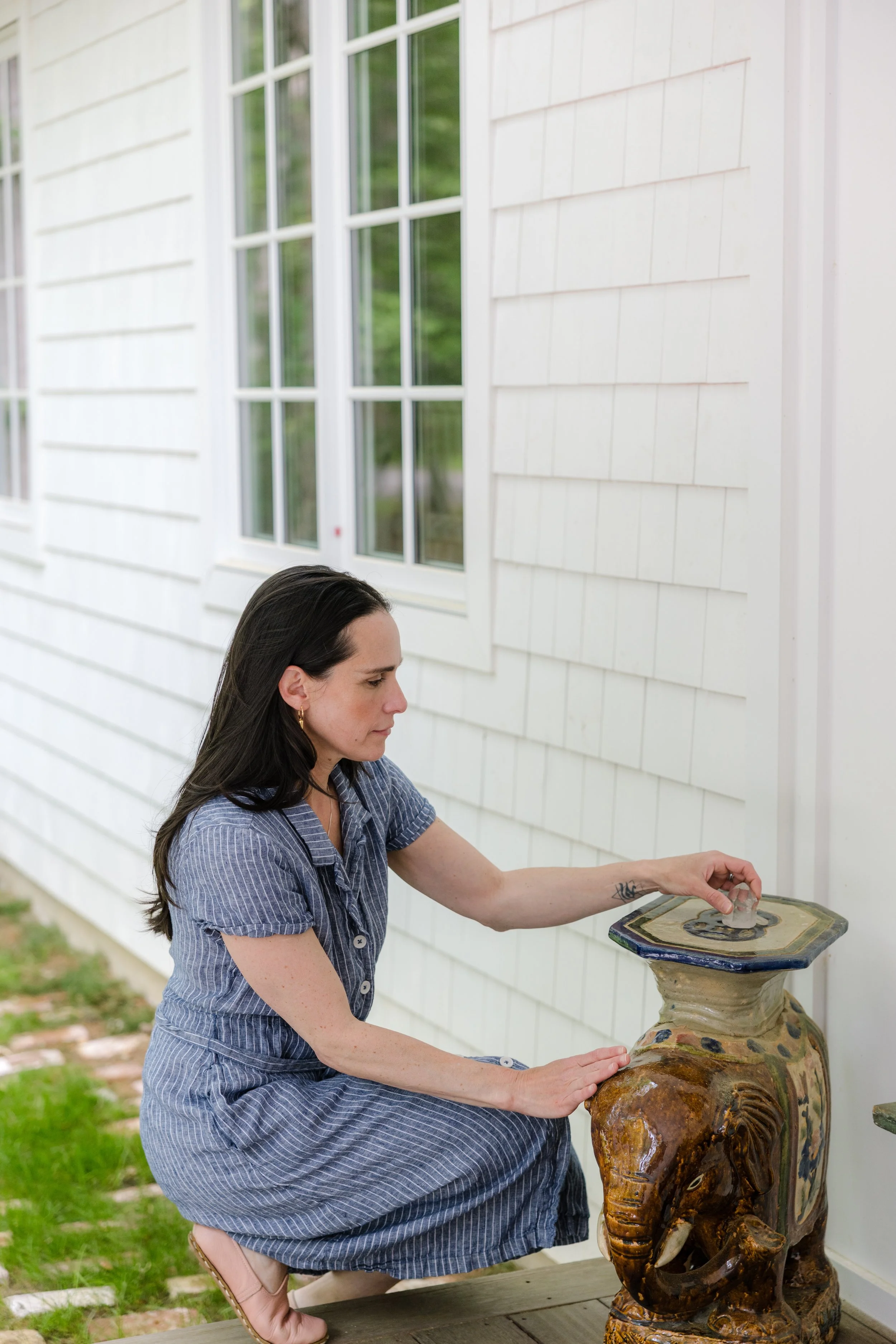A woman in a blue striped dress is outside, placing a coin into a large, decorative ceramic elephant statue used as a birdbath or fountain, against the backdrop of a white house with large windows.