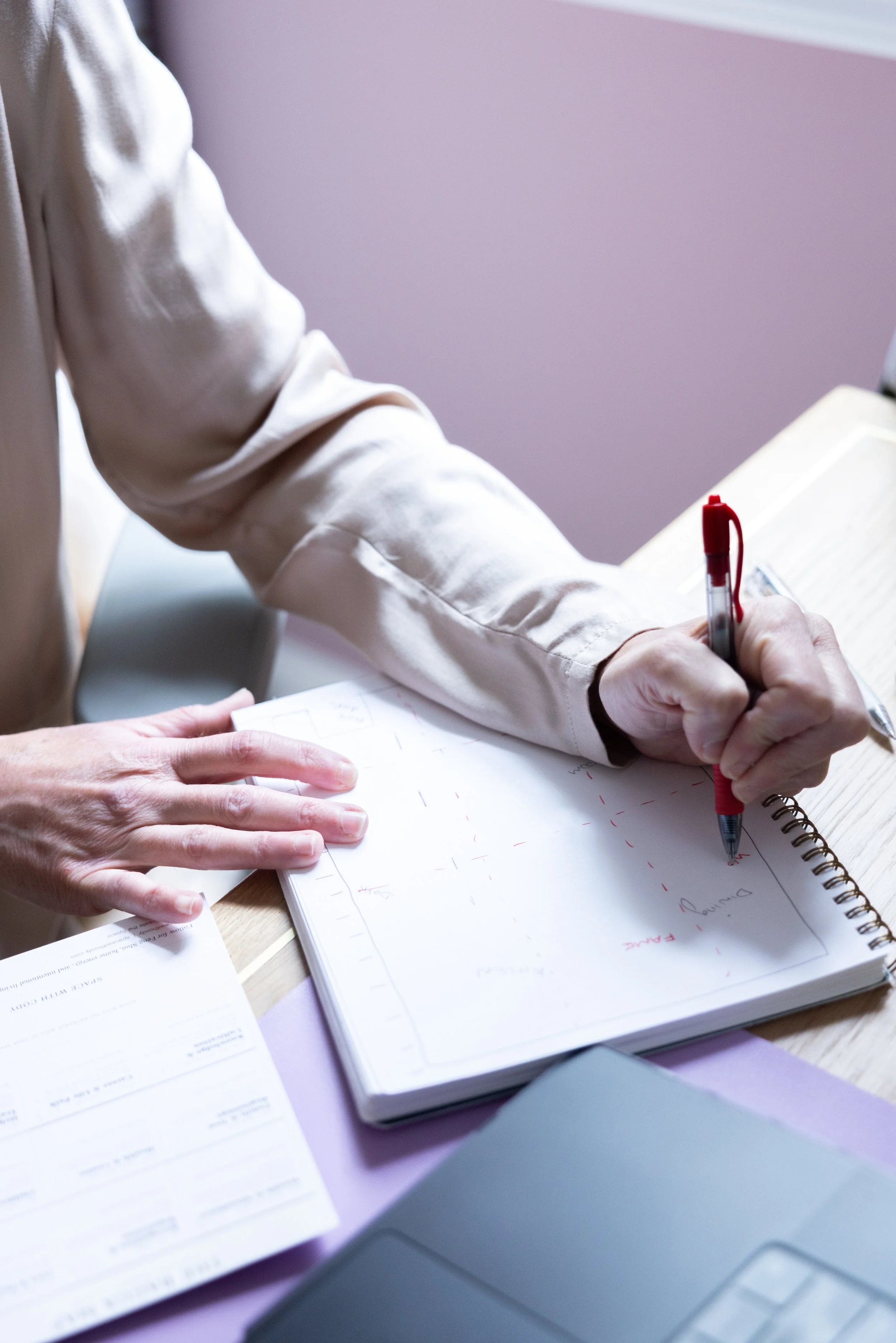 A person wearing a beige sweater writing in a notebook with a red pen at a wooden desk. There is a paper with printed text and a laptop nearby.
