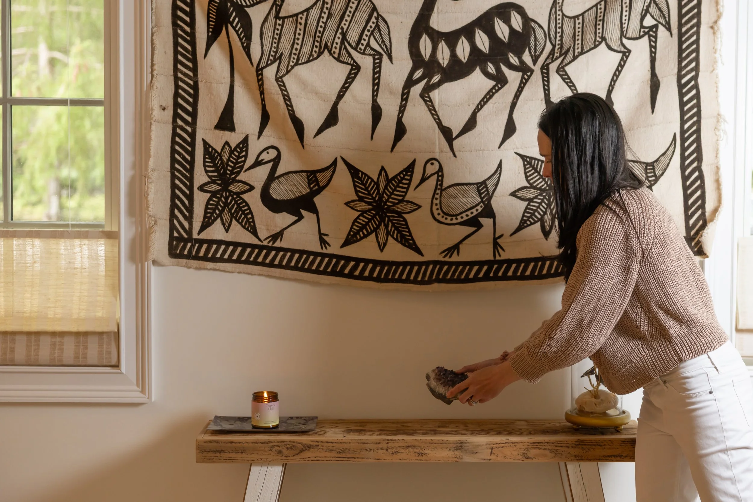 Woman with dark hair and light sweater in a room with a large black and white tribal-style wall hanging featuring animals and leaves, a window with bamboo shades, a candle on a table, and a decorative orb on a side table.